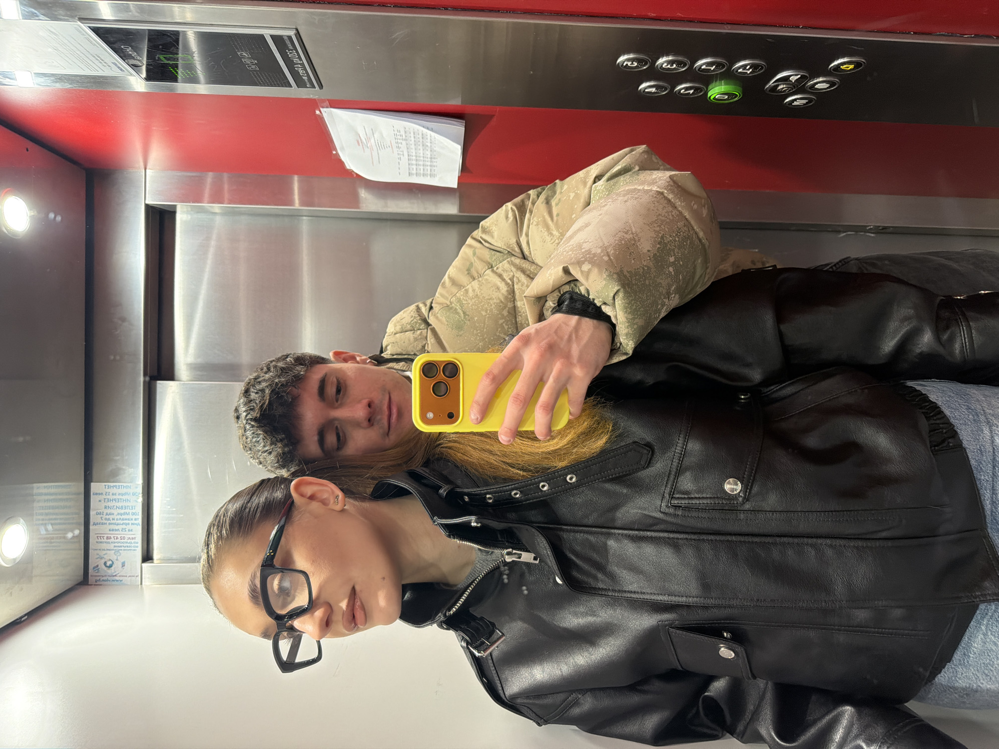 Two people, a man and a woman, are taking a selfie in an elevator. The woman, wearing eyeglasses and a black leather jacket, is in the foreground. Behind her lies the man, wearing a camouflage puffer jacket. They are holding a yellow smartphone to take the picture. The elevator has a red and silver interior. There's a panel with elevator buttons. There are signs on the walls. The visible text on one sign appears to be written in Russian. The image was most likely taken in Sofia, Bulgaria.