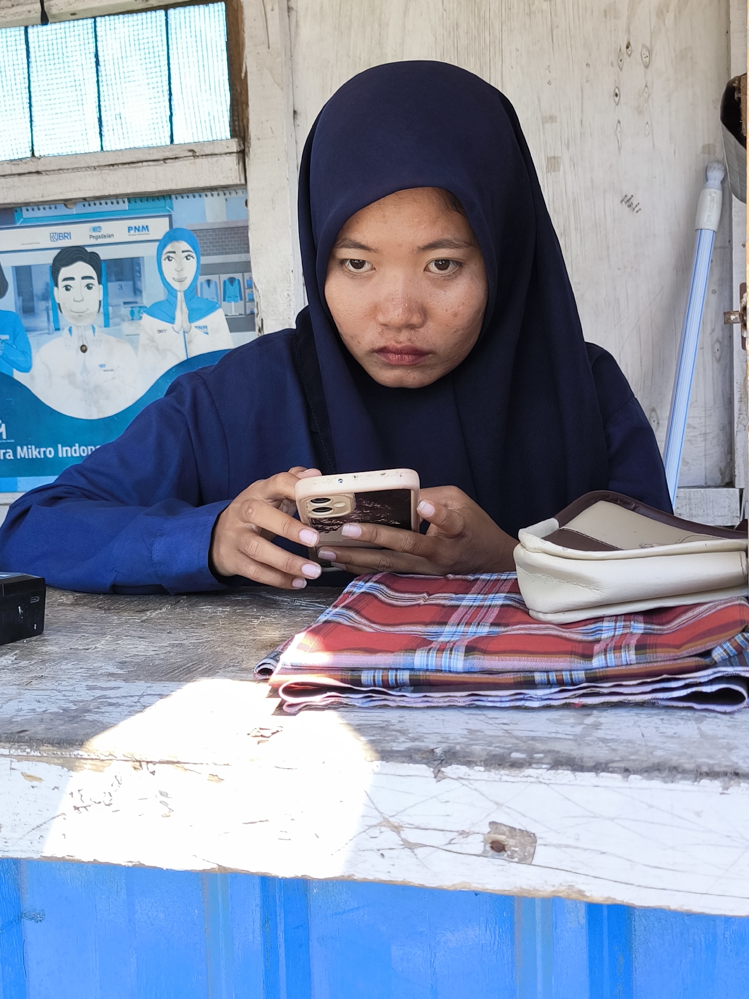 A woman wearing a dark blue hijab is engrossed in her smartphone, resting her elbows on a worn table. A folded plaid cloth and a small purse are positioned on the table next to her. Behind her, a poster featuring cartoon figures is visible. A blue-handled cleaning tool leans against the wall. The image was captured in Labuhan Lombok, Indonesia.