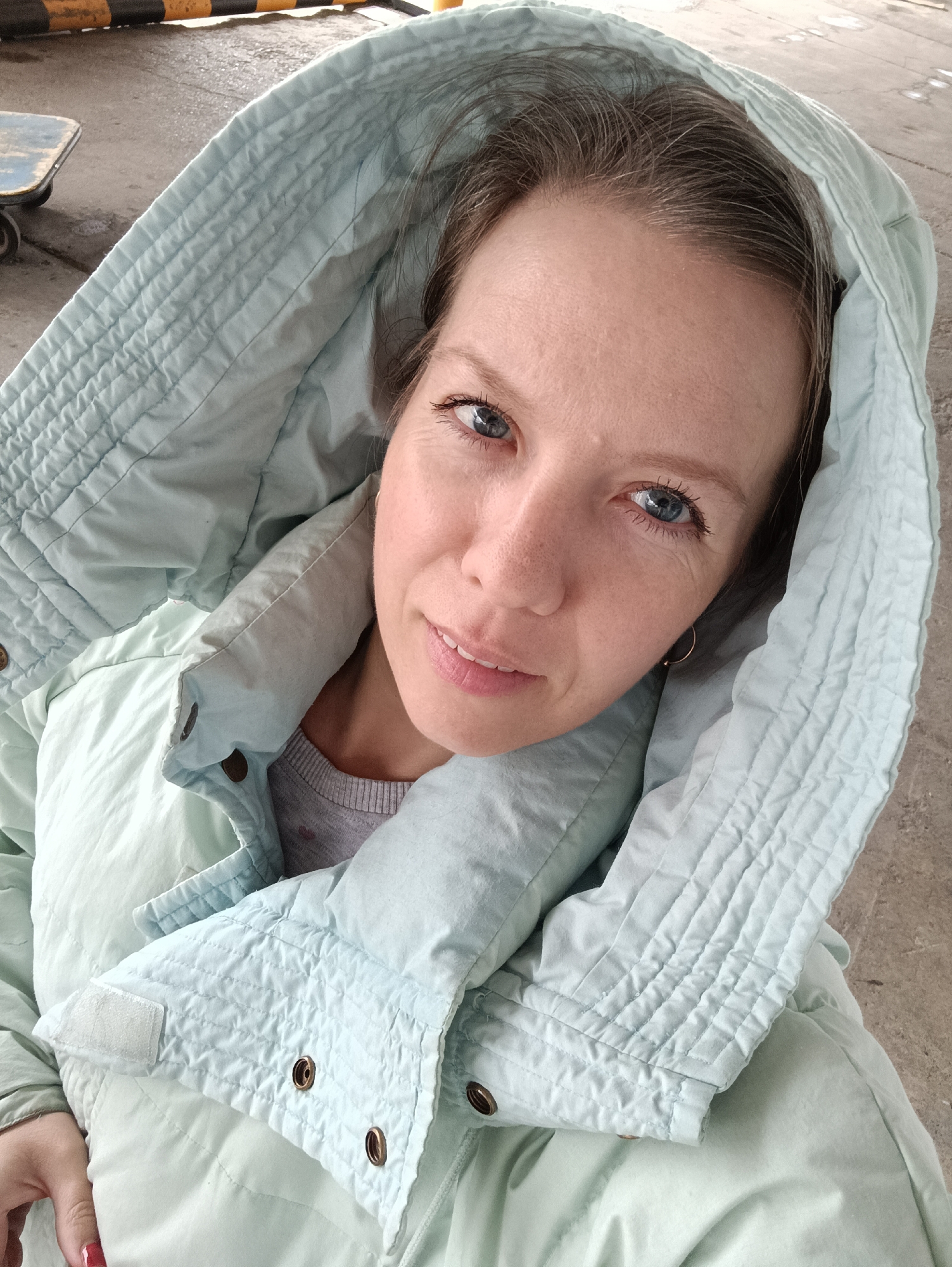 A close-up, high-angle portrait features a woman looking directly at the camera, likely a selfie. She is wearing a light blue quilted jacket with the hood up, framing her face. A gray long-sleeved shirt is visible underneath the jacket. Her blue eyes are prominent, and she displays a subtle, pleasant smile. Her hair is brown with noticeable gray strands, particularly at the temples. A small gold hoop earring is visible in her left ear. Her hand is partially visible at the bottom left, showing a fingernail with dark red polish.
The background is an outdoor or semi-outdoor setting in Chaikovsky, Russia. Elements in the background include concrete ground, a portion of a blue and white skateboard with wheels, and a yellow and black striped barrier or object. The lighting is bright and diffused, characteristic of daytime, possibly an overcast day or a shaded location. The quilted jacket suggests cool weather. There is no visible text in the image. The overall impression is casual and direct.