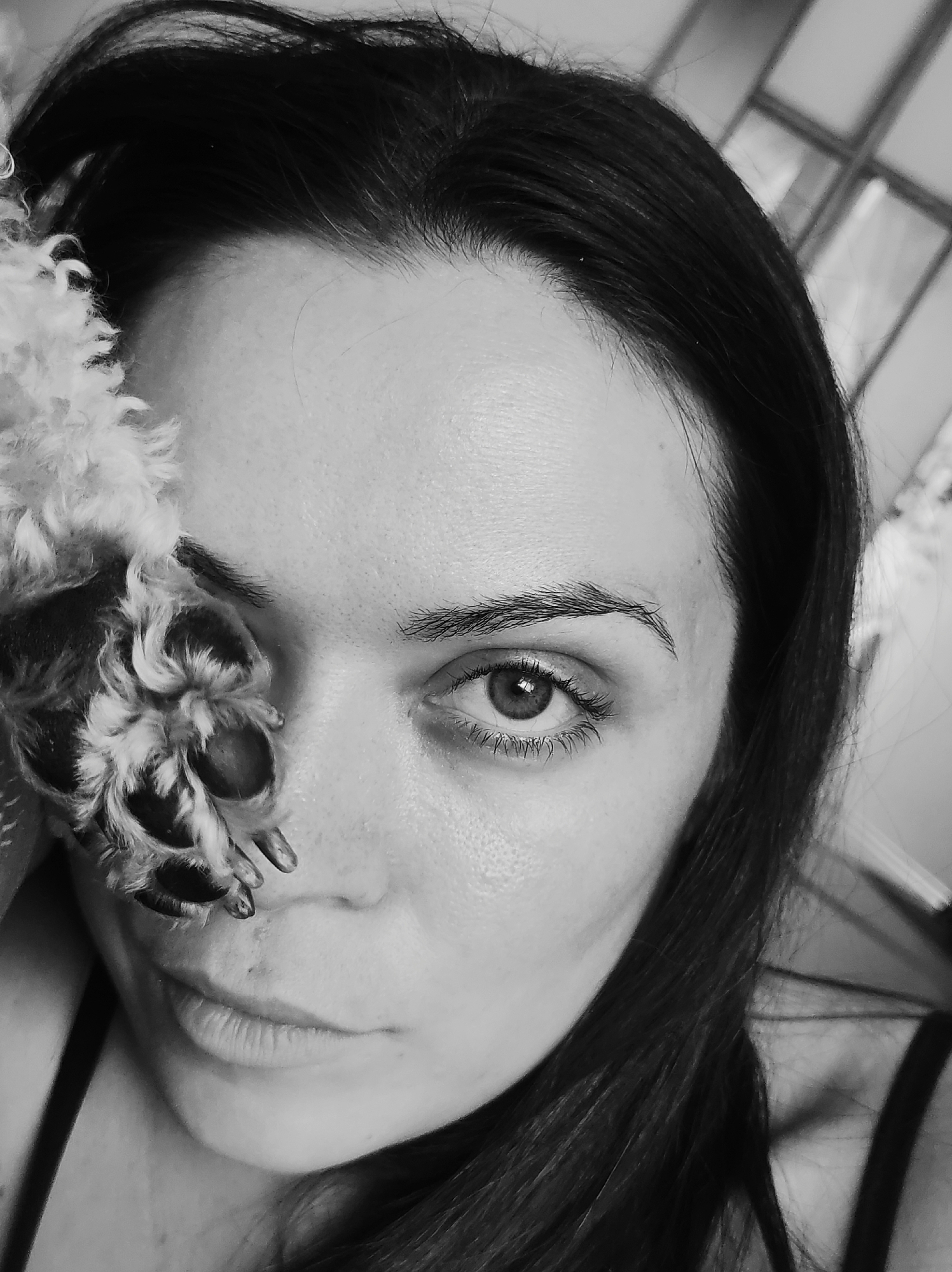 The image is a close-up black and white photograph of a woman's face, partially obscured by what appears to be a dog's paw. The woman has dark hair, fair skin, and one visible eye. Her expression is neutral. A dog's paw, covered in fur and with exposed claws, is positioned over part of her face, near her eye. The background is slightly blurred. The location of the image is not apparent from the provided context or the cropped image set. It is not possible to determine if this image was taken in Chaikovsky, Russia.