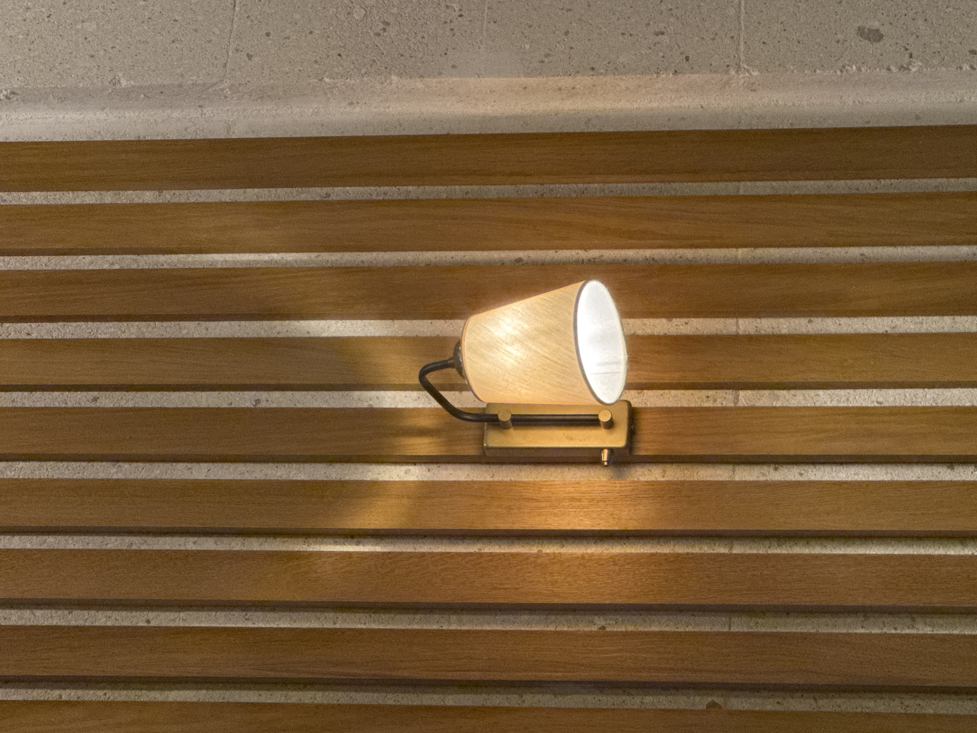 A wall sconce illuminates a wall with alternating horizontal bands of brown wood and light gray stone. The sconce has a conical shade of light beige material with a black gooseneck arm and a rectangular bronze base. The wood bands are evenly spaced and display a subtle grain. The stone bands appear rough and textured. The lighting from the sconce casts a warm glow. The location is not evident from the image.