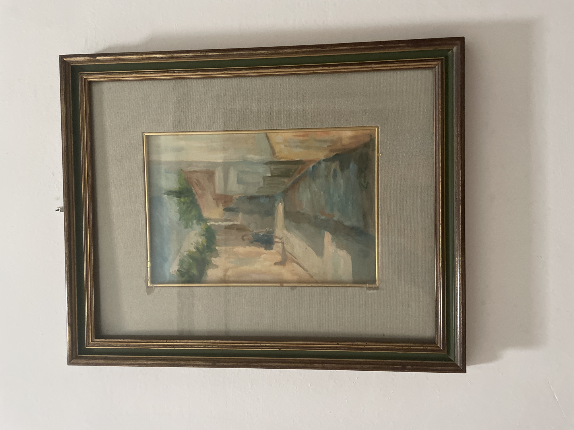 A framed painting of a narrow street scene hangs on a white wall. The painting depicts a cobblestone street lined with buildings. A single figure, possibly a child in a dark dress, walks away from the viewer down the street, casting a shadow. The sky above is a light blue with hints of clouds. The painting is matted with a light gray fabric and framed with a dark wood frame with a green inset.