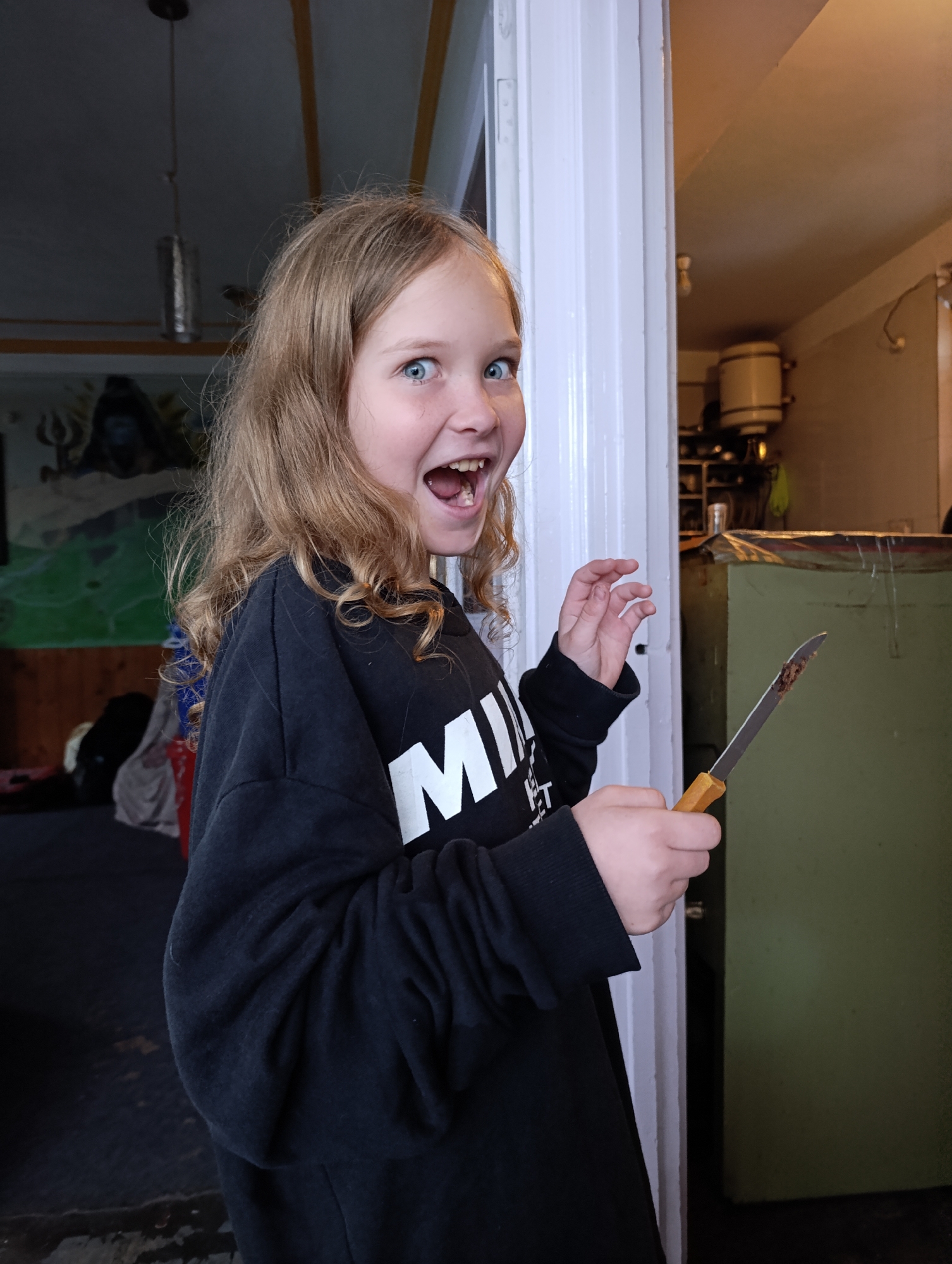 A young girl with blonde, curly hair is standing in what appears to be a doorway, possibly in Vashisht, India, given the context. She is wearing a black sweatshirt with white lettering. Her mouth is open in a surprised or excited expression, and her eyes are wide. She is holding a knife with a brown handle in her right hand, and there is some dark material on the blade. Her left hand is raised with fingers splayed. The background is somewhat blurred, showing a dimly lit room with what looks like a painting on the wall and some industrial-looking equipment.