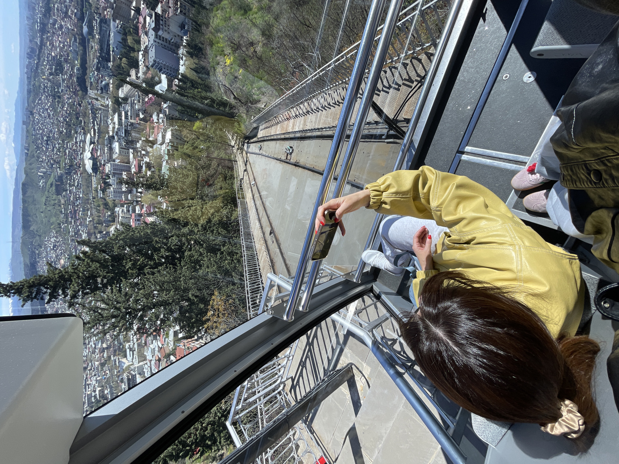 A person wearing a mustard-colored jacket, gray pants, and pink shoes is sitting inside a funicular in Tbilisi, Georgia, holding a black phone. The funicular track and a cityscape are visible outside the window. The person has dark hair with a scrunchie.