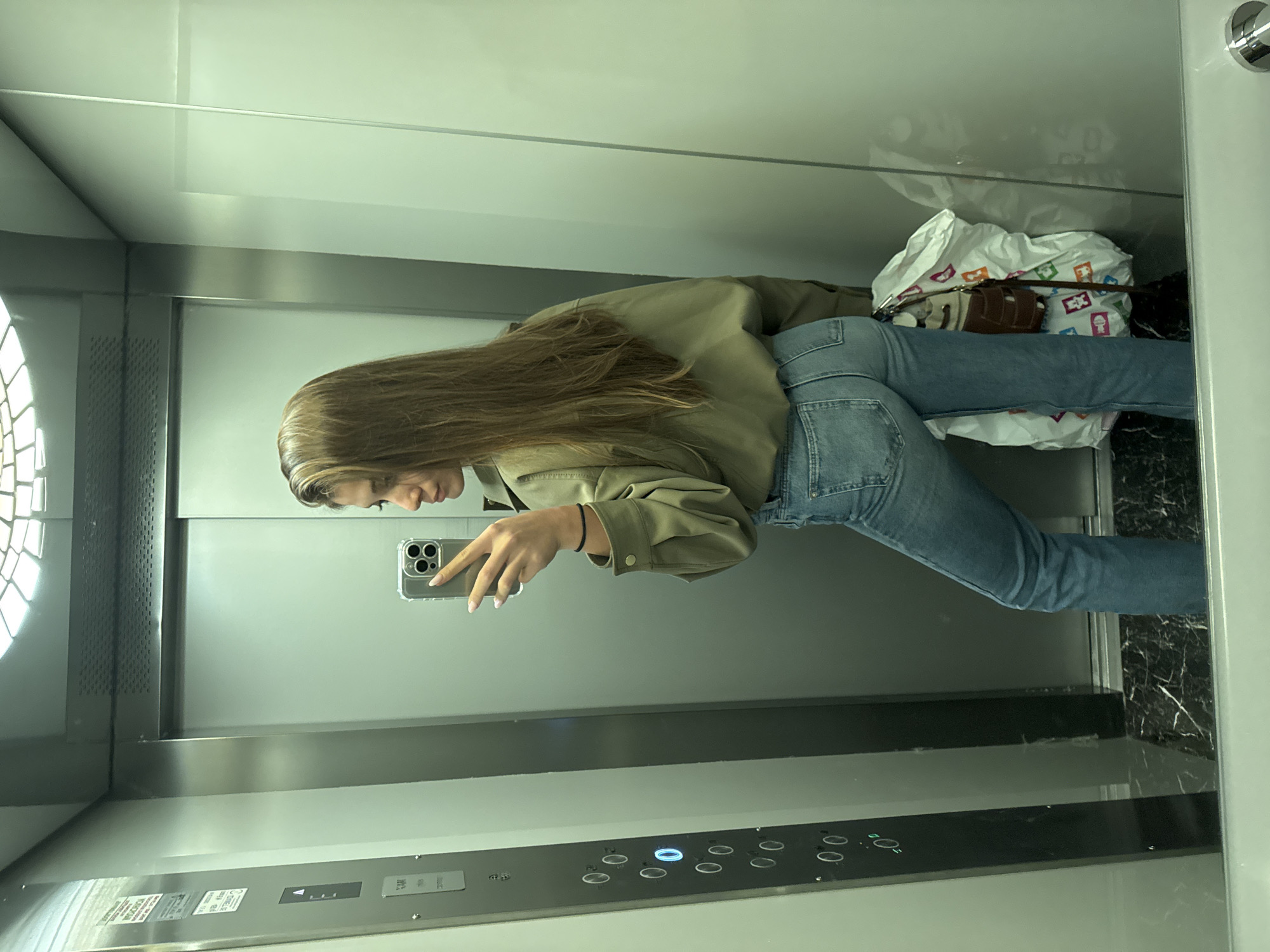 A young woman is taking a mirror selfie in an elevator. She is wearing jeans and an olive green jacket. She is holding a smartphone in her right hand. Behind her are bags and what appears to be a purse. The elevator has a panel of buttons. The location in the city of Burgas, Bulgaria is not discernible from this image.