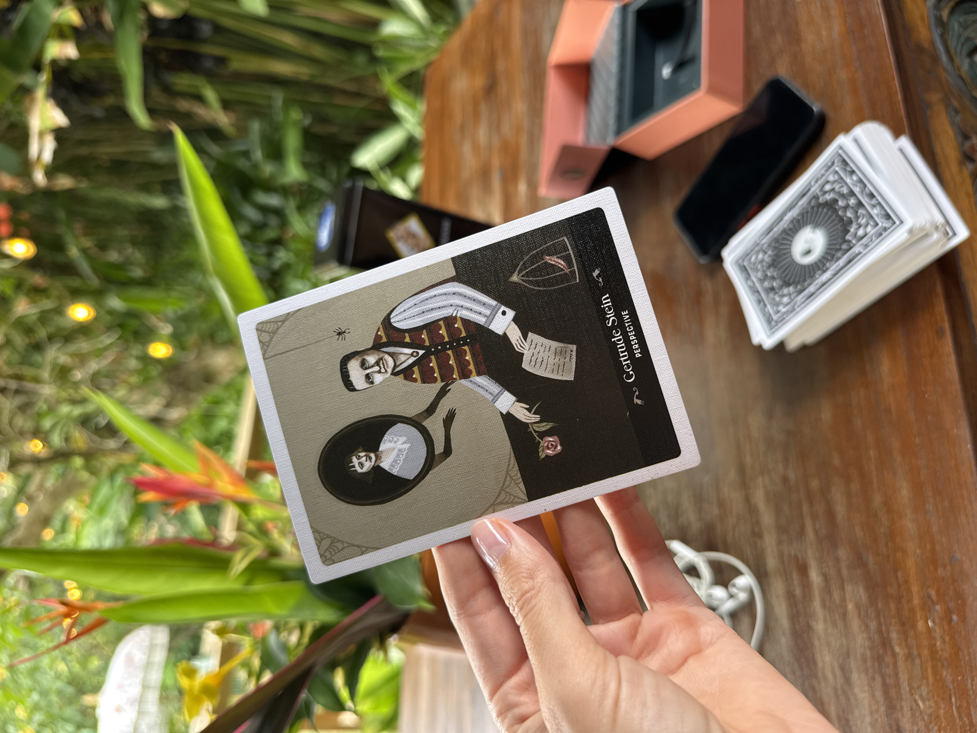 A person's hand with a manicured fingernail is holding up a tarot card featuring an illustration of Gertrude Stein and a woman in a portrait. The card reads "Gertrude Stein PERSPECTIVE." The card is placed on a wooden table in what appears to be an outdoor setting with lush green foliage and tropical flowers in the background. Also on the table are a stack of cards, a cell phone, and an open coral-colored box. The setting is likely a cafe or resort in the city of Mengwi, Indonesia, given the tropical flora.