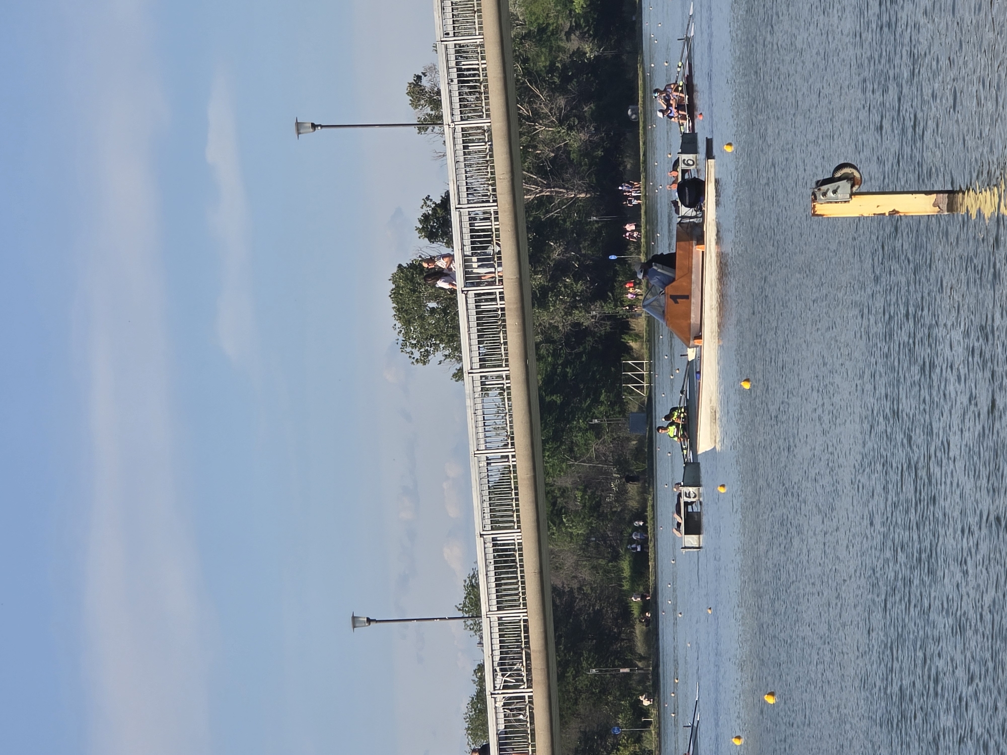 The image depicts a rowing competition on a body of water, possibly a river or lake, in an urban or suburban setting. Several rowing teams are visible, each in their respective lanes marked by yellow buoys. The teams are composed of multiple rowers in long, slender boats.
A pedestrian bridge runs alongside the water, with a few people visible on it observing the race. The presence of streetlights along the bridge further indicates an urban location. The environment includes trees lining the side of the water and a clear sky in the background, suggesting a sunny day. A starting platform or dock is positioned on the water, with the number "1" visible on its side. A yellow metal structure is located near the platform.
The precise location in the city of Unknown, Unknown, cannot be determined without further information or landmarks.