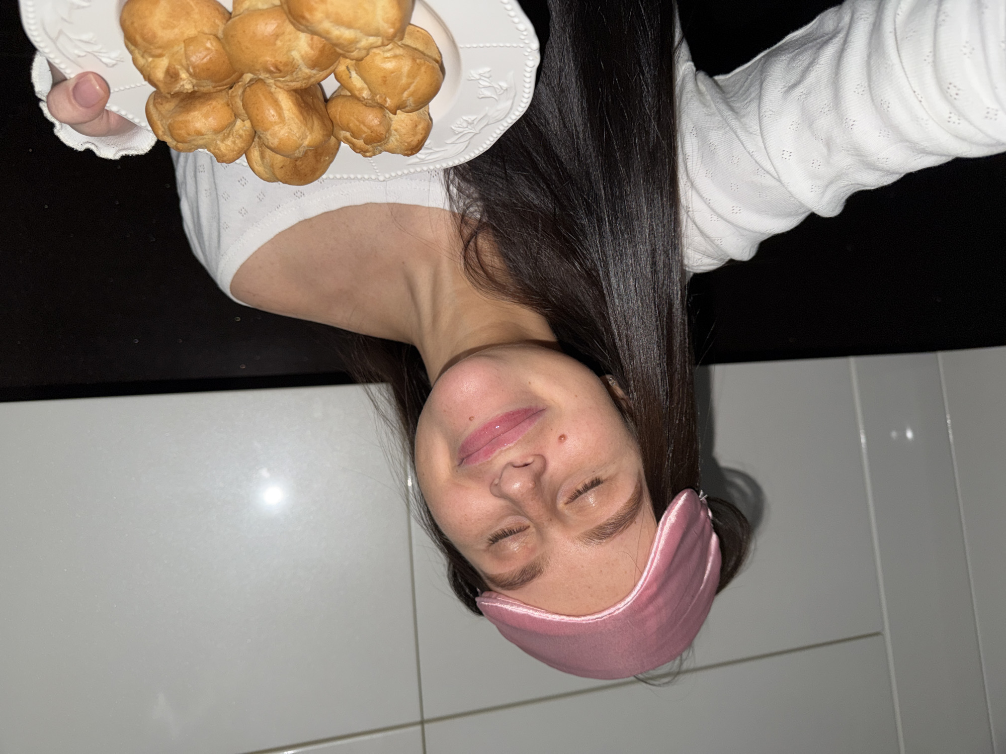 A young woman with long dark hair is holding a white plate with a mound of golden-brown pastries, possibly profiteroles or cream puffs. She wears a white long-sleeved shirt and a pink satin sleep mask pushed up on her forehead. She has her eyes closed and a slight smile. The background includes a dark surface (likely a countertop) behind the plate and white tiled walls. The location is not discernable from the image and cannot be confirmed to be in Khimki, Russia.