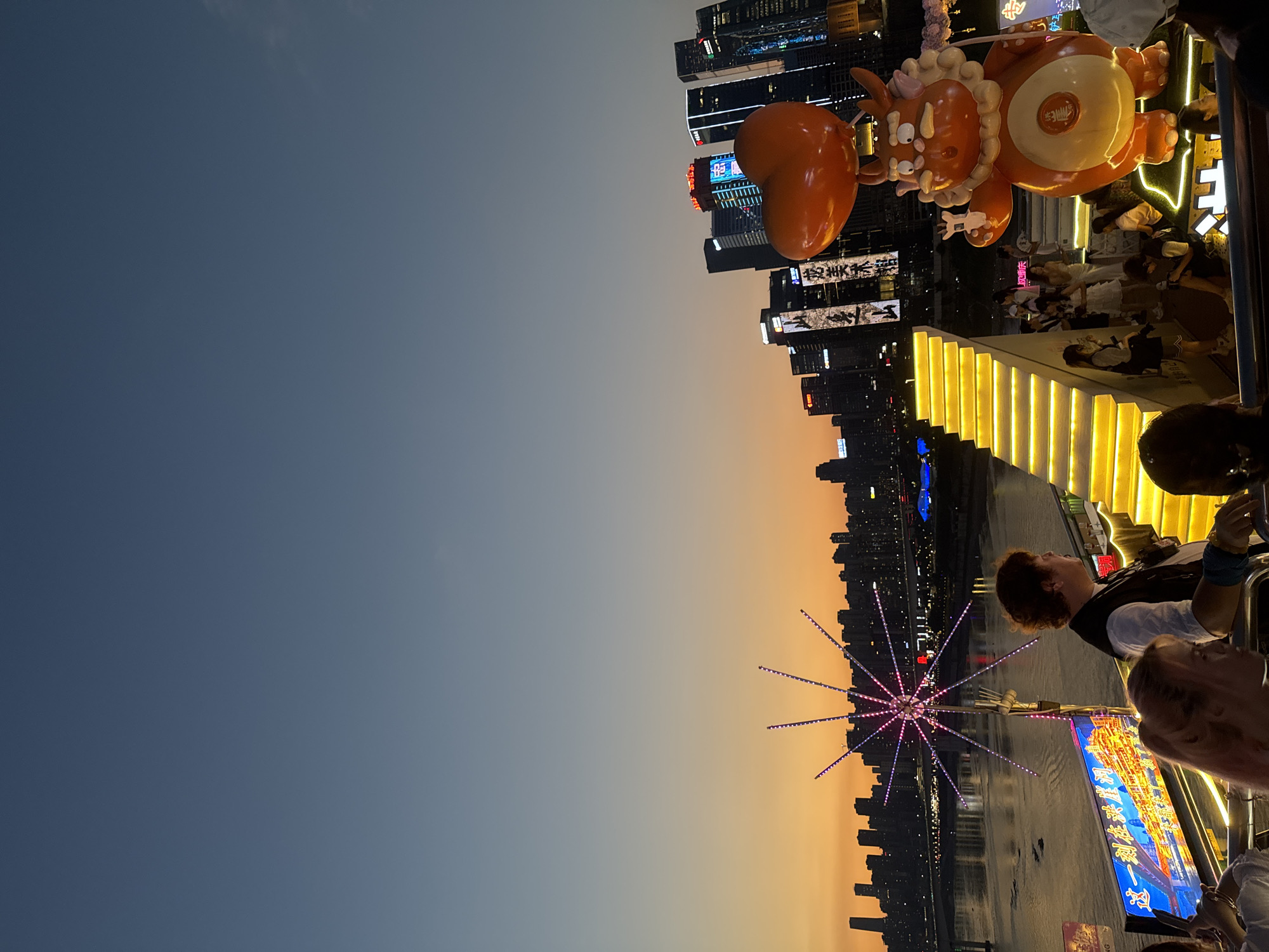 The image captures a vibrant waterfront scene at dusk in Yuzhong District, China. In the foreground, a large, orange, cartoonish dragon-like mascot holds a heart-shaped balloon. To its left, a set of brightly lit, yellow stairs ascend. Several people are visible on and around the stairs, some looking out at the cityscape. In the mid-ground, a star-shaped light installation with pink and purple lights stands on a platform overlooking a river. A billboard with Chinese characters and an image of a fantastical city is mounted below it. Across the river, the Yuzhong District skyline is illuminated with numerous tall buildings and bright lights, reflecting on the water's surface. The sky exhibits a gradient of deep blue at the top, transitioning to warm orange and yellow hues near the horizon, indicating the setting sun.