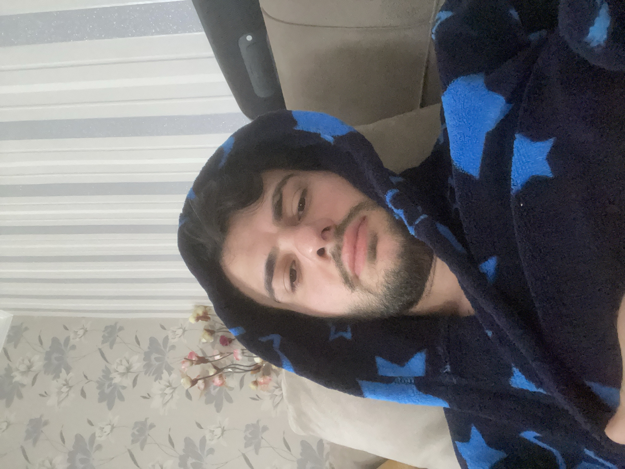 A man is lying on a couch, wearing a dark blue hooded robe with light blue star patterns. He has dark hair and a beard. He appears relaxed, looking at the camera. The couch is light brown. The background includes a wall with a floral pattern and a slatted window shade. The specific location within Tbilisi, Georgia cannot be determined from the image.