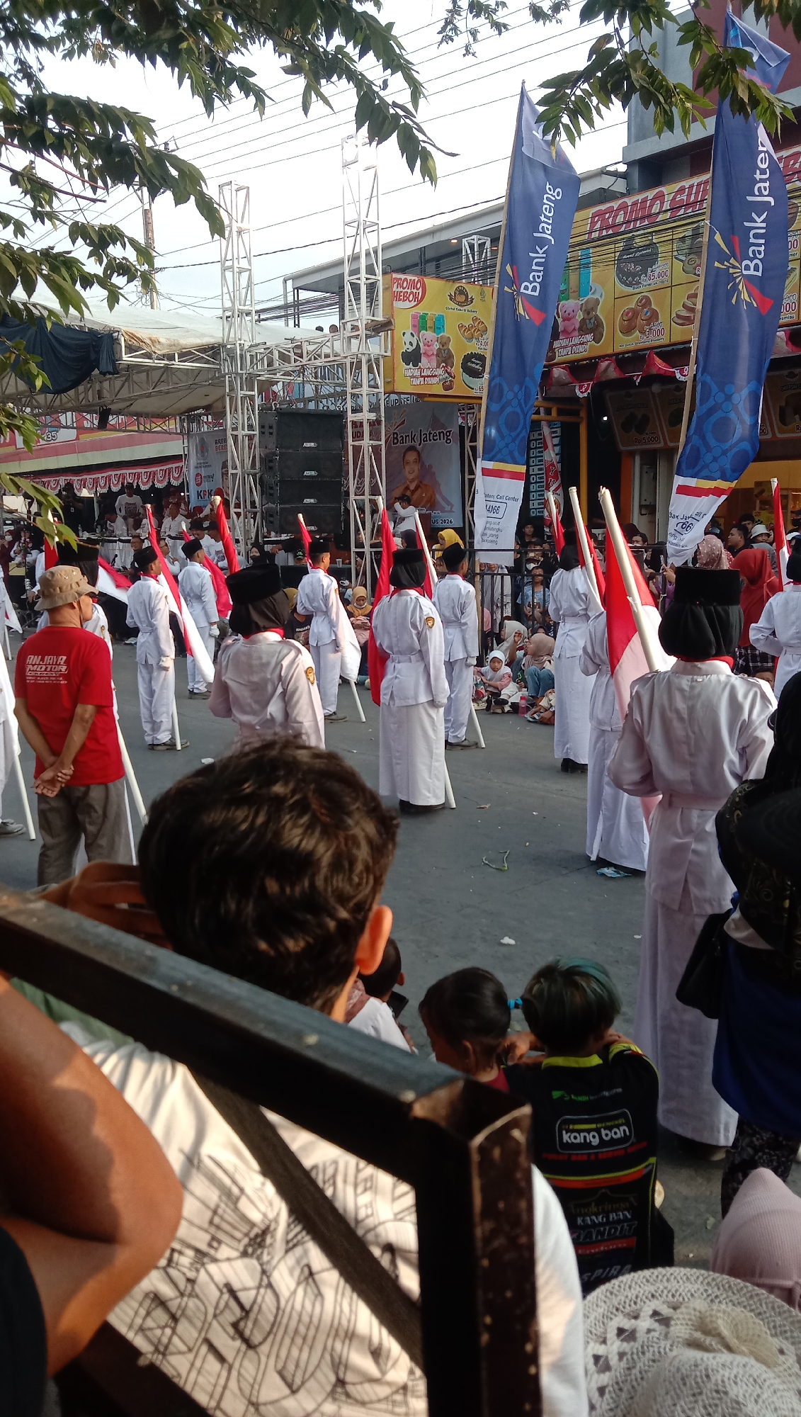 This is an outdoor, eye-level, medium shot captured on a sunny day, possibly in Jatisawit, Indonesia, depicting a parade or performance. In the foreground, the back of a person's head and shoulder, wearing a white shirt with an abstract design, are visible, along with a dark wooden railing. The middle ground features a group of people dressed in white uniforms and black hats, holding red and white flags, likely participating in a ceremonial event. Some of these individuals appear to be marching or standing at attention. Behind them, a man in a red shirt and a hat stands with his hands clasped behind his back. Further in the background, a stage with large speakers and scaffolding is set up, adorned with banners and a large image of a person. There are also spectators in the background, including children sitting on the ground. The environment appears to be a street or open area, with buildings and trees visible in the distance. The overall atmosphere seems festive, with the flags and uniformed participants suggesting a patriotic or cultural celebration.