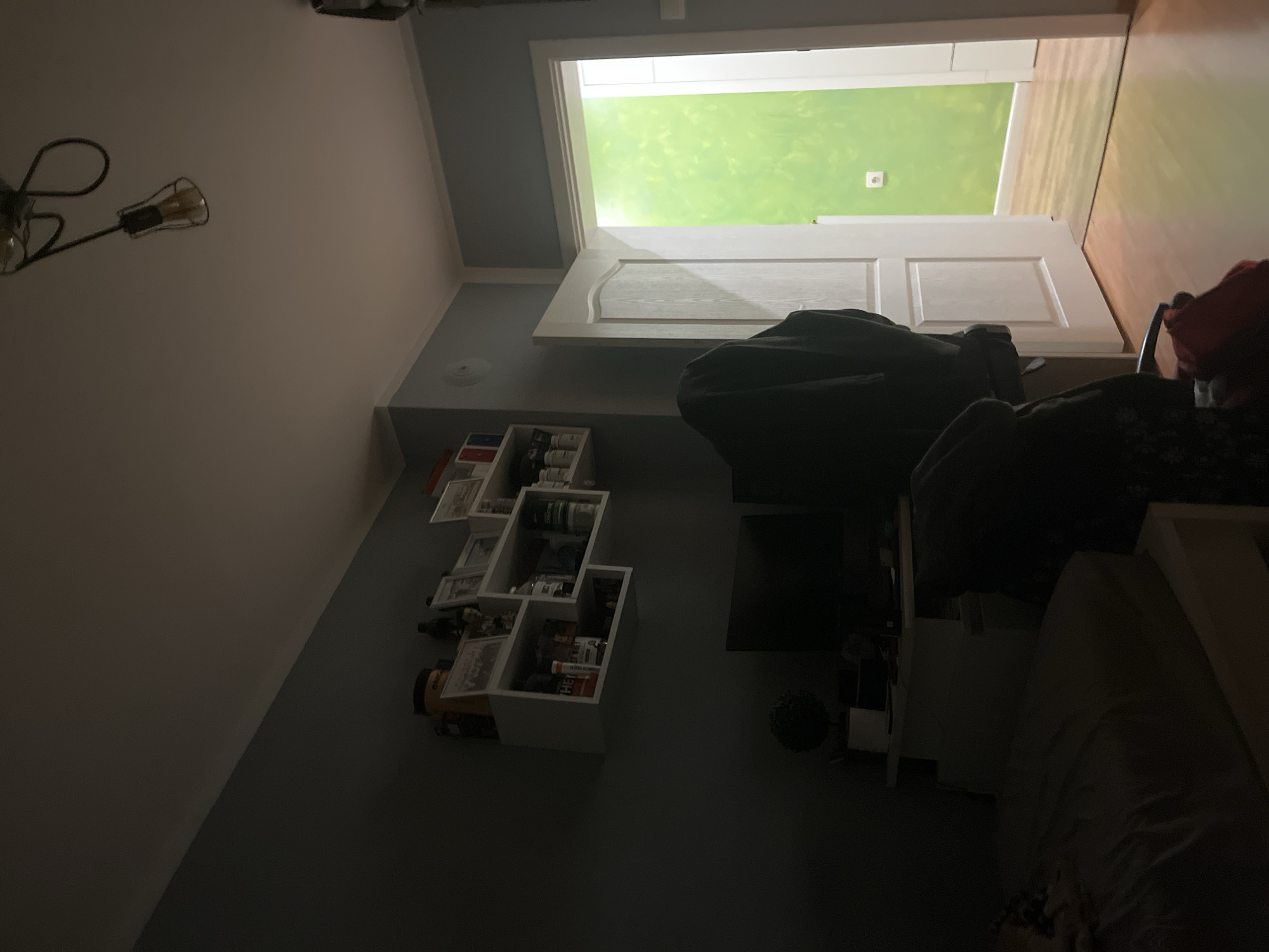 The image shows an interior view of a room. The walls are painted in two shades, a light color near the ceiling and a darker shade below. There's a light fixture with a retro-style bulb hanging from the ceiling. A white, irregularly shaped shelving unit is mounted on the darker wall, filled with various items. A door is ajar, leading to a room painted in green. The floor is visible, made of light-colored wood. In the foreground, there's a bed with dark bedding, along with some dark-colored furniture and other objects. The scene is dimly lit. There is no information about the city of Pokrovnik, Bulgaria within the image.