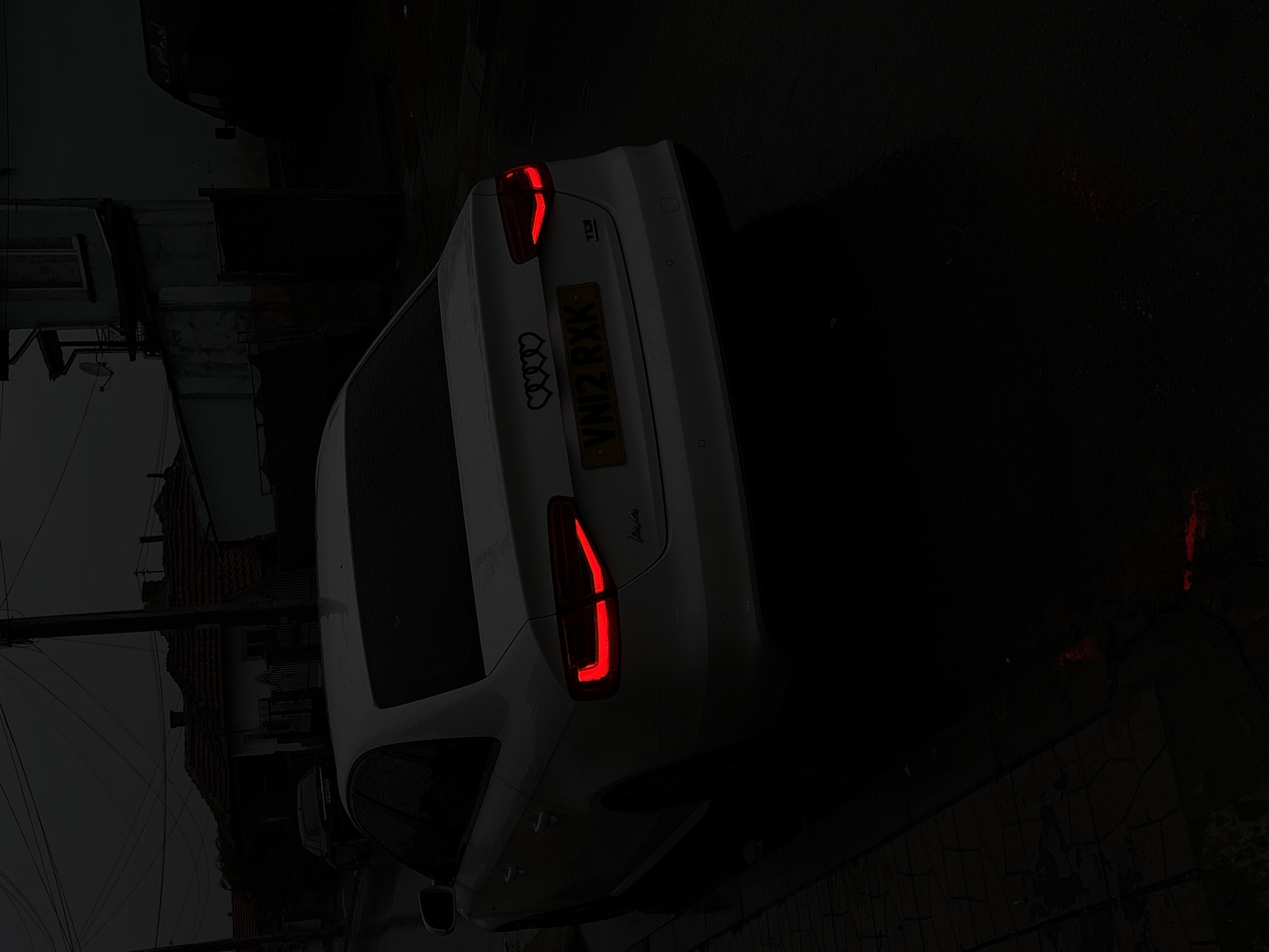 The image shows the rear of a white Audi A4 parked on a street in Dolni Tsibar, Bulgaria. The vehicle's taillights are illuminated. The registration plate is visible and reads VN12 RXK. The surrounding environment appears dark and wet. Buildings are visible in the background, but details are obscured by the darkness. A utility pole is present on the left side of the image. The focus is primarily on the car and its features.