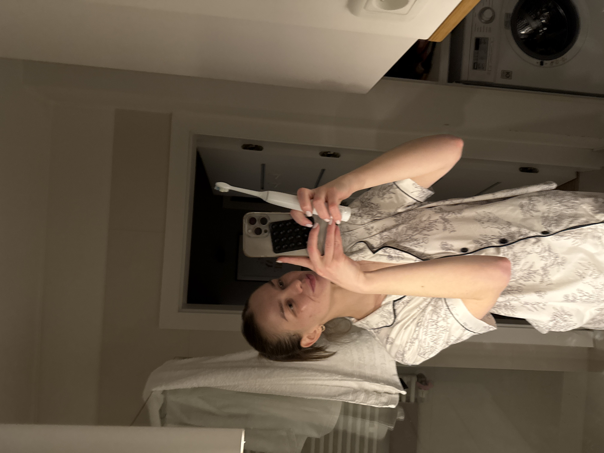 A woman in patterned pajamas is holding a smartphone and a white electric toothbrush in a bathroom. The woman is standing in front of a mirror with a white towel draped over the edge. Behind her is a window, a washing machine, and cabinets. The location is a bathroom in an unknown location within Warsaw, Poland.