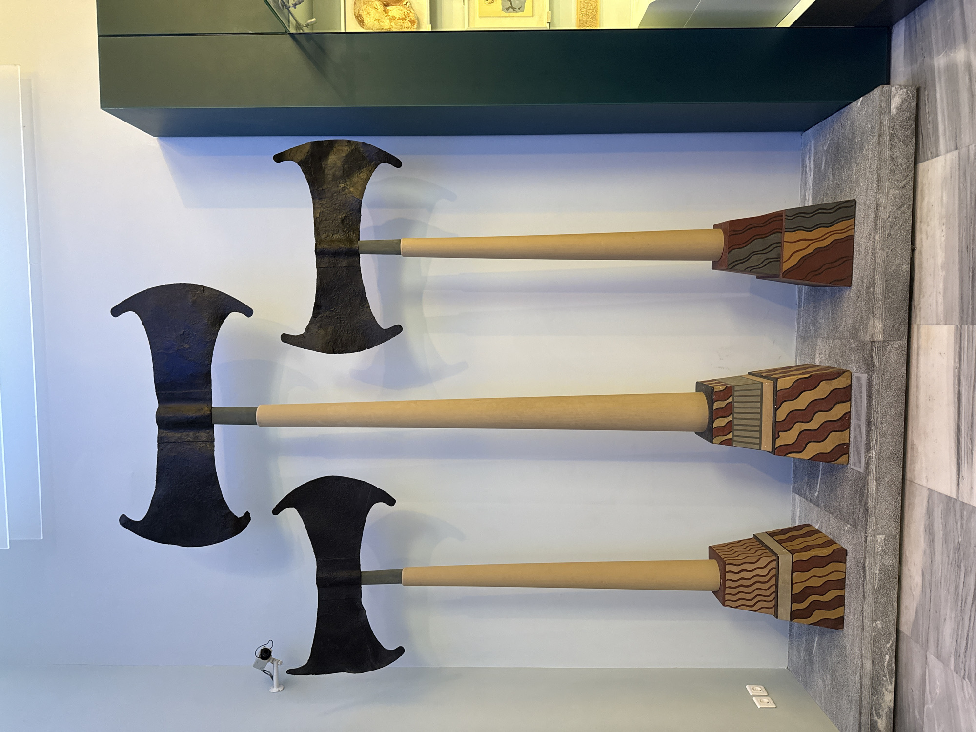 In Heraklion, Greece, within a museum setting, three imposing double-bladed axes are displayed. The axes, each with a dark, textured blade, are mounted on light-colored, cylindrical shafts. These shafts rise from decorative, tiered bases that feature geometric patterns in brown, red, and yellow. The axes are arranged from left to right in decreasing height, with the tallest in the center. They stand on a grey, stone platform that extends across the foreground. The background is a plain, light blue wall, with a security camera visible on the left. To the right, a dark green display cabinet with glass fronts offers a glimpse of artifacts within, including what appears to be pottery or stone objects. The floor is tiled with a marble-like pattern.