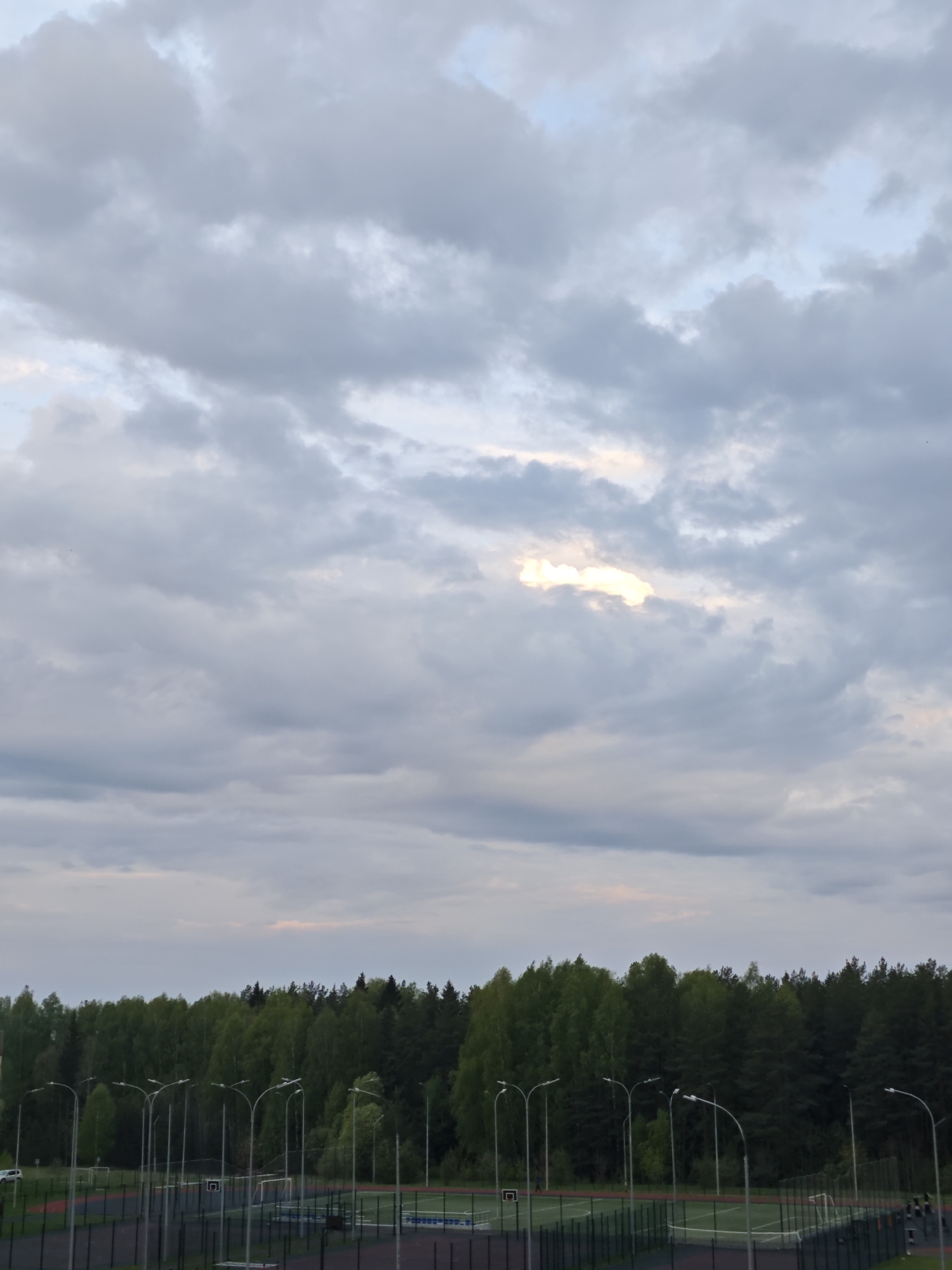 The image depicts an outdoor sports complex in Markovskii, Russia, viewed from an elevated position. A forest borders the complex. Metal fencing surrounds the area. Several curved streetlights are present, illuminating a running track and various athletic fields. Overcast skies dominate the upper portion of the image, with a single patch of sunlight visible through the clouds.