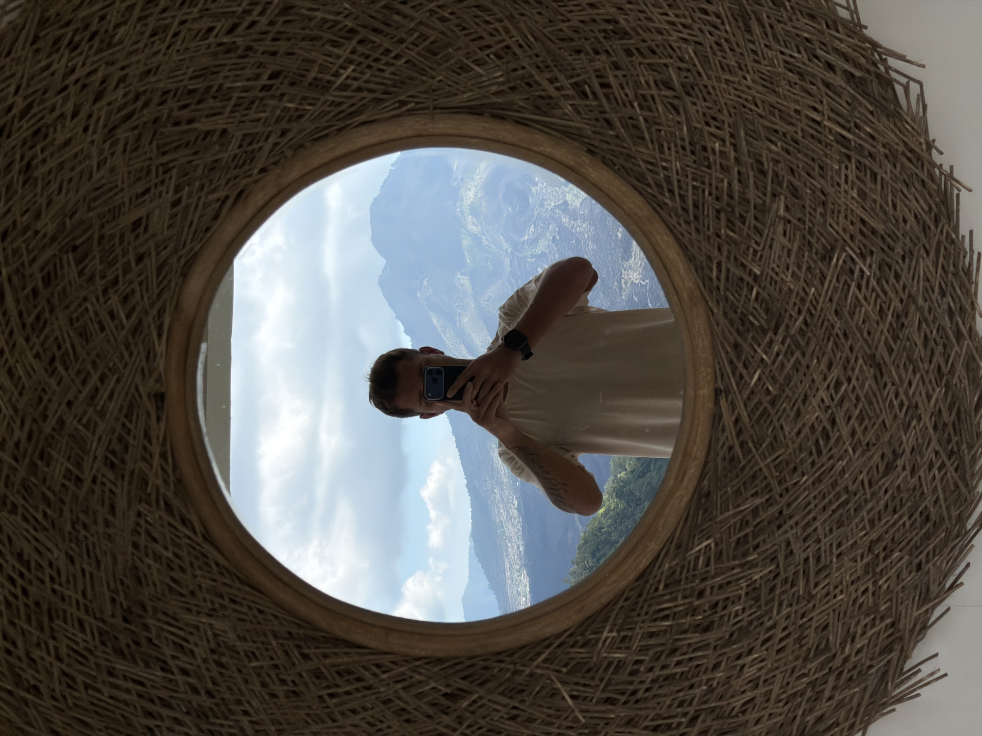 A man stands in front of a mirror, taking a selfie. He is wearing a light-colored t-shirt and a watch on his left wrist. The mirror is framed with woven material and reflects a mountainous landscape with a cloudy sky. The man is holding a smartphone in his right hand, with his left hand extended upwards to capture the photo. The reflection shows the man's torso and arms. In the background, visible through the mirror, are rolling hills and distant mountains, suggesting an outdoor or scenic location, possibly in Kintamani, Indonesia, given the mountainous terrain. The overall impression is a casual self-portrait set against a natural backdrop.