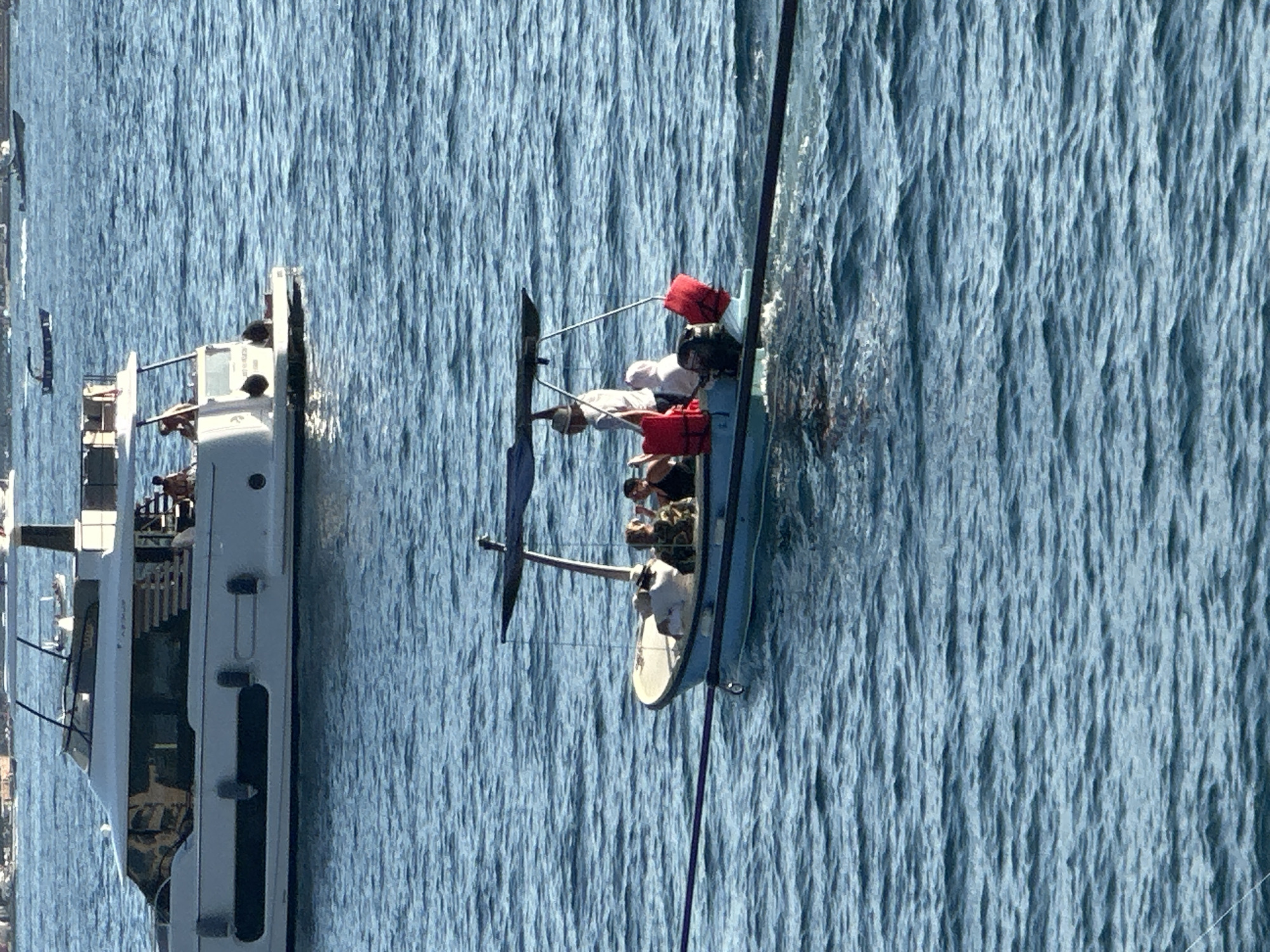 A wide shot captures a bright, sunny day at sea, likely near Budva, Montenegro, given the presence of both a large white yacht and a smaller blue boat. The sea is a vibrant blue with gentle waves. In the foreground, the smaller boat, approximately 5-7 meters in length, carries four individuals. Two red life vests are clearly visible, one attached to the back of the boat and another on a seat. The boat has a canopy for shade, supported by metal poles. A person in a light-colored shirt and hat stands near the rear of the boat, while others are seated. The larger white yacht, in the background, is significantly larger, with multiple levels and windows, suggesting it is a luxury vessel. Several people are visible on the upper deck of the yacht. The overall scene depicts a leisurely day on the water, with two distinct types of watercraft present.