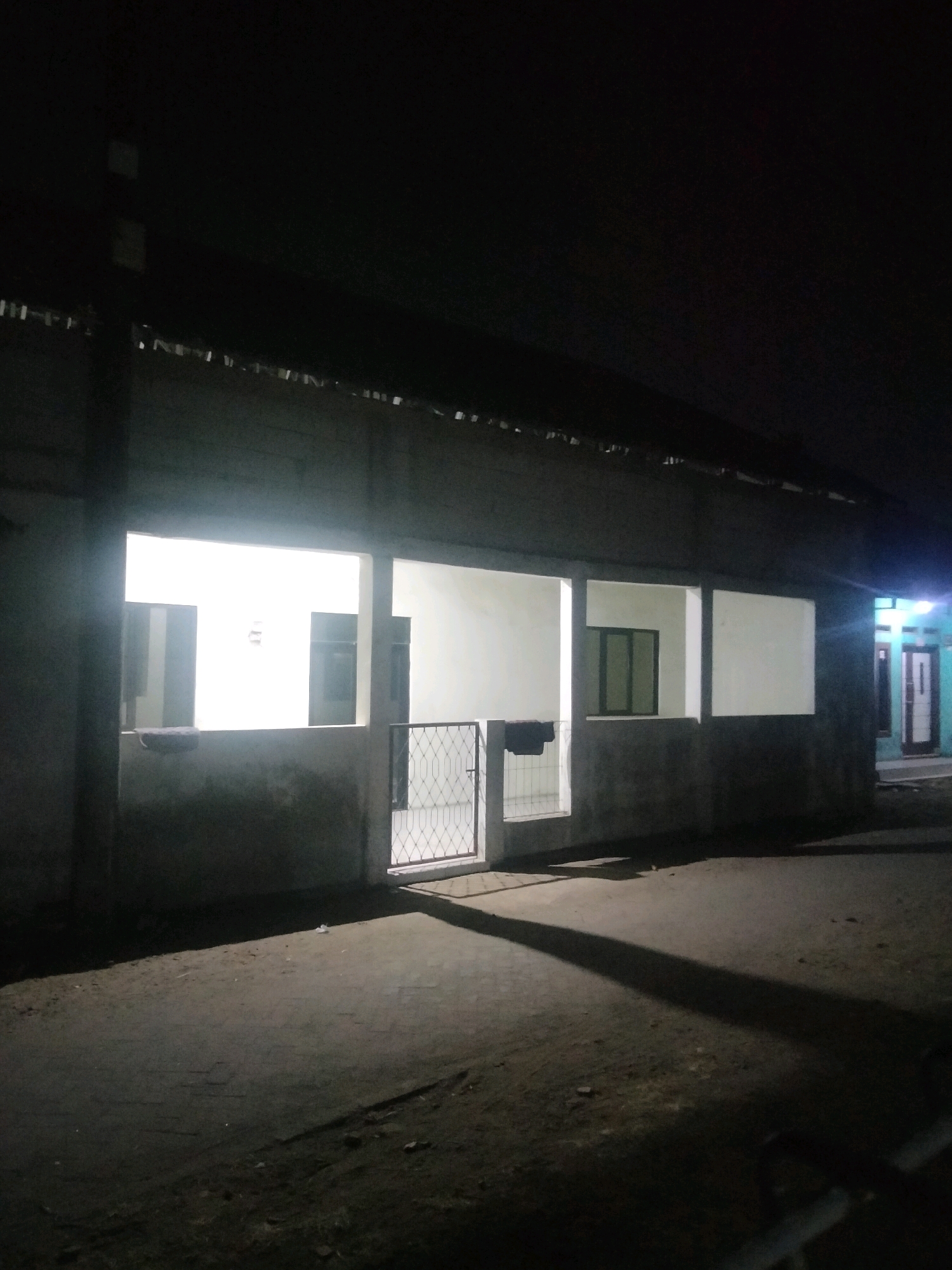 The image depicts a nighttime view of a building exterior in the city of Suka Sari, Indonesia. The building appears to be a single-story structure with a covered walkway or porch along the front. Several large openings, possibly windows or doorways, are visible. The interior of some of these openings is brightly illuminated, suggesting lights are on inside. One opening on the left has a dark object resting on its ledge, which could be a bag or blanket. To the right of this opening, a metal gate with a diamond pattern is visible. Further to the right, another opening shows a window with green panes. In the distance, a doorway is illuminated by blue and white lights. The ground in front of the building is paved with a dark material, possibly asphalt or concrete, and is partially covered in dirt and debris. A long, distinct shadow stretches across the ground from the left side of the image towards the right, indicating a light source outside the frame. The overall lighting suggests it is after dark, with artificial lights illuminating the scene.