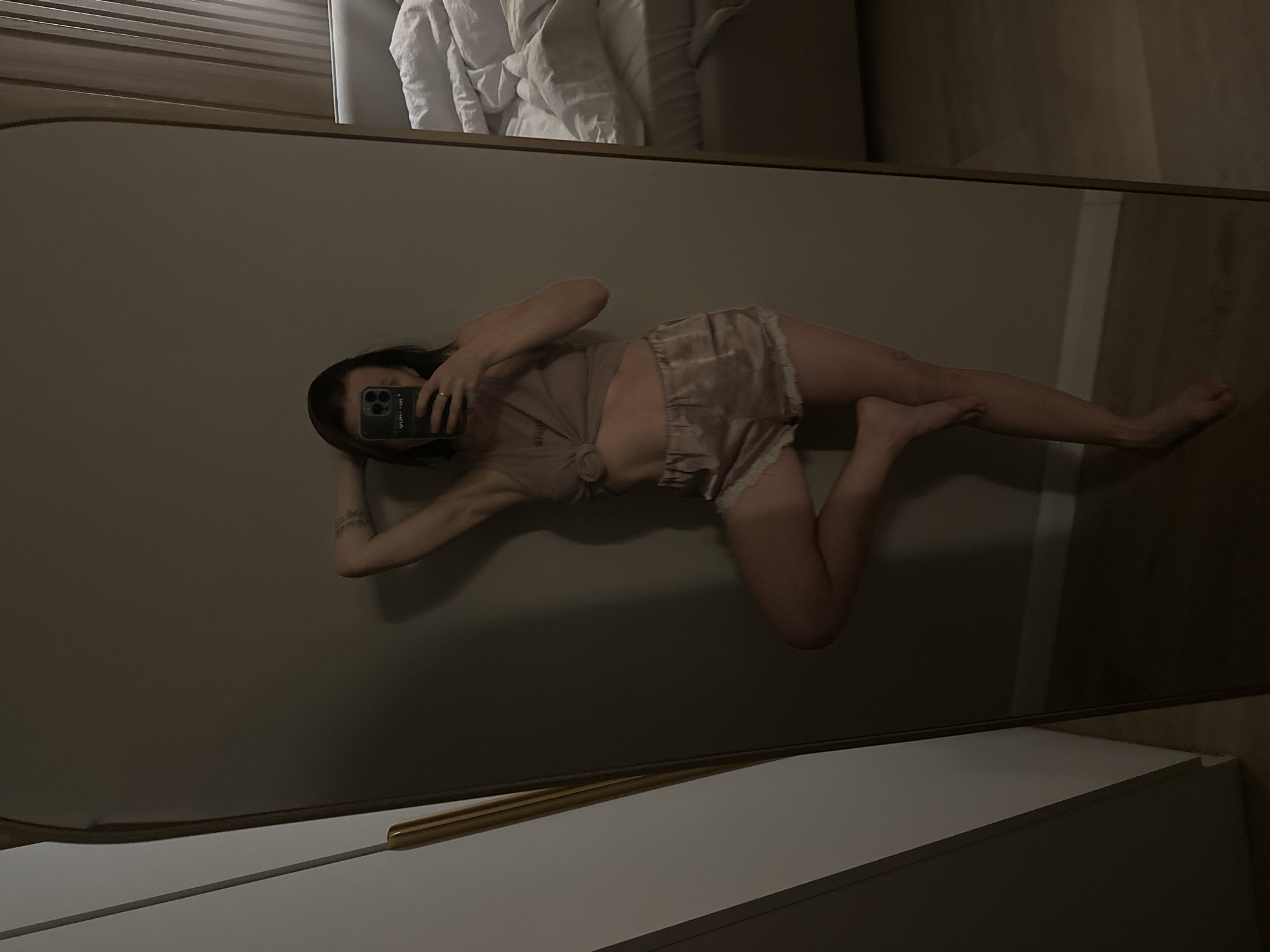 A person lies on their side, seemingly posing for a mirror selfie. They are wearing a neutral-colored top and shorts. The mirror's reflection includes a bed with white bedding and a dresser. The location is indiscernible from the image.
