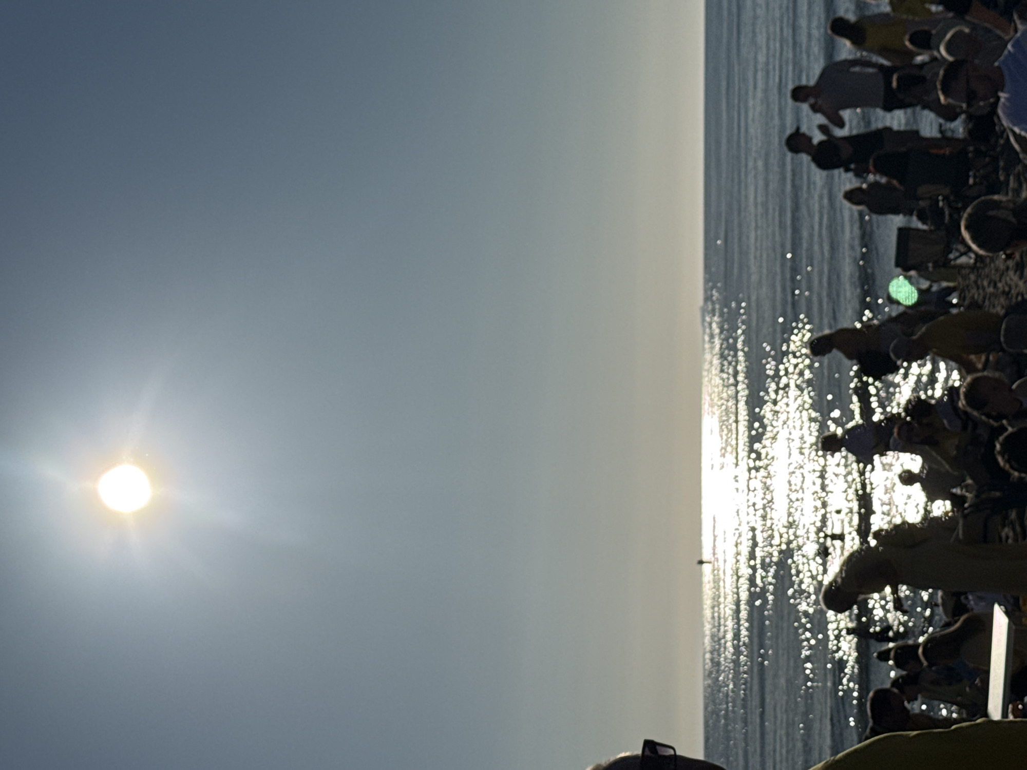 The image depicts a beach scene with a bright sun shining in the upper left quadrant of the frame. The sun is casting a strong glare across the water, creating a glittering effect on the surface. A hazy horizon separates the sky from the ocean. In the foreground, silhouetted figures of people are visible on the sand, some standing, some sitting, and some in motion. They appear to be gathered in a casual setting. A few individuals are in the process of walking or interacting with each other. The beach itself is sandy, and the water appears calm. There is a person in the distance standing on a small pier or buoy.
