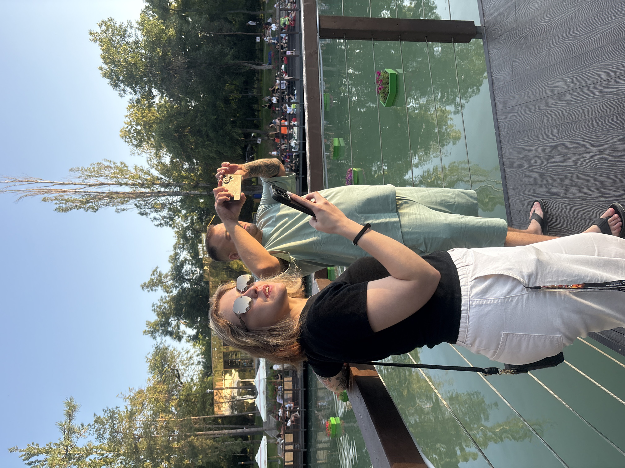Two people are on a wooden deck overlooking a body of water. The person in the foreground, a woman with blonde hair, wears a black t-shirt and white pants. She is wearing sunglasses and looking upwards with a smile, suggesting a relaxed and pleasant mood. The second person, a man behind her, wears a light green shirt and is holding a smartphone, seemingly taking a photo. He has tattoos on his arm. The scene appears to be outdoors on a sunny day, with trees and a clear blue sky visible in the background. The water reflects the surrounding trees and sky. In the distance, there is a gathering of people sitting at tables under umbrellas, indicating a public or recreational area, possibly a cafe or park. The presence of decorative green planters with flowers floating in the water adds to the scenic ambiance. The overall impression is one of a leisurely outing during daytime, possibly in Almaty, Kazakhstan, given the provided location context.