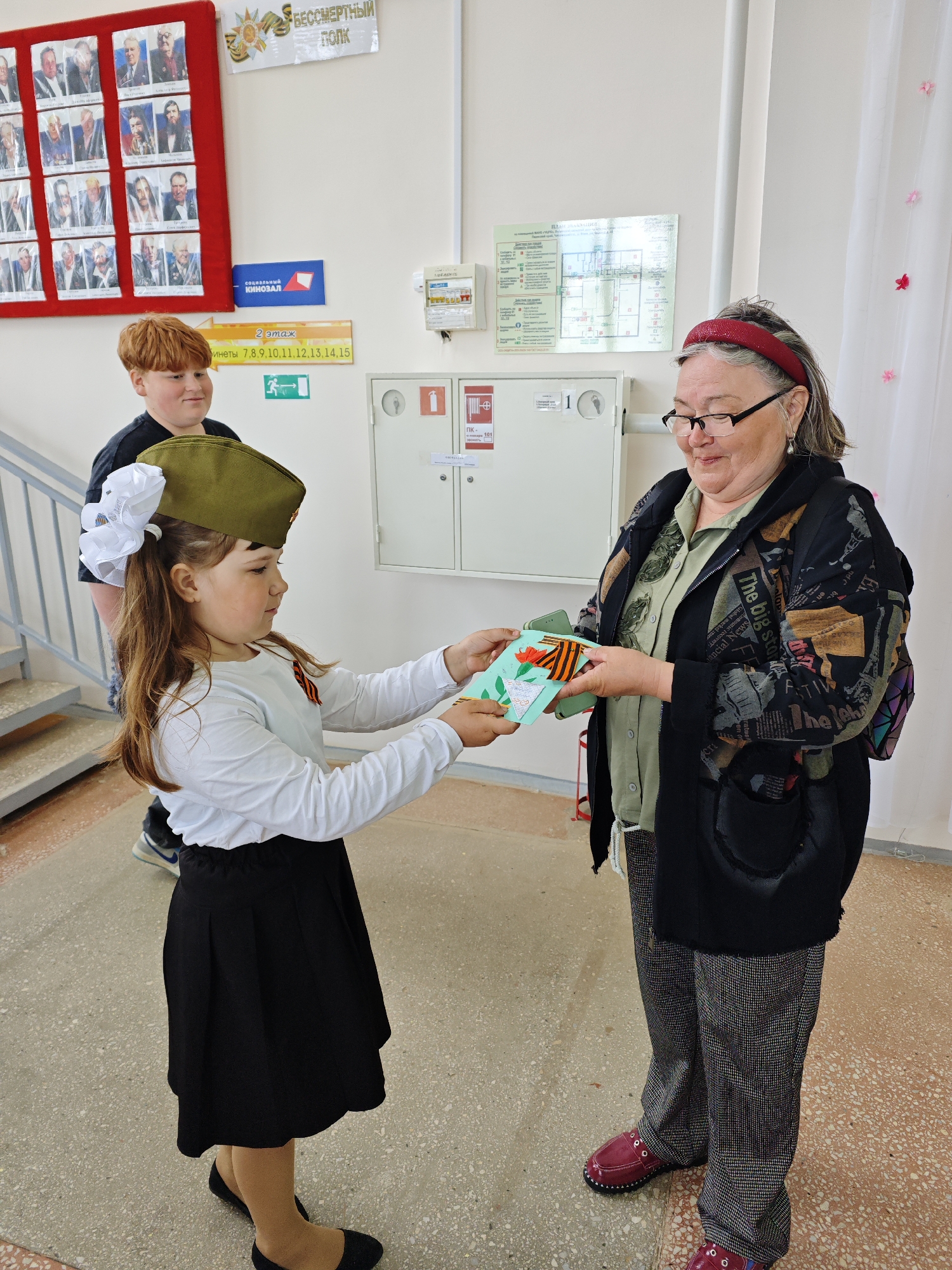 A young girl presents a gift to an older woman indoors, likely in a school. The girl wears a black skirt, white long-sleeved shirt, black shoes, and a military-style hat adorned with a white bow. She holds a small, decorated box with a ribbon. The woman, wearing glasses, a printed black jacket over a green shirt, and patterned pants with red shoes, receives the gift. Behind the girl stands a redhead boy in a black t-shirt. The setting appears to be a school hallway, with a bulletin board, fire safety equipment, a staircase, and signage in Russian visible in the background. A sign that says "КИНОЗАЛ" (cinema hall) and lists room numbers suggests a cinema or auditorium area. There's also a poster indicating it is a "Бессмертный Полк" (Immortal Regiment) event, commonly associated with Victory Day commemorations in Russia. This indicates that this image was taken in Фоки, Russia.