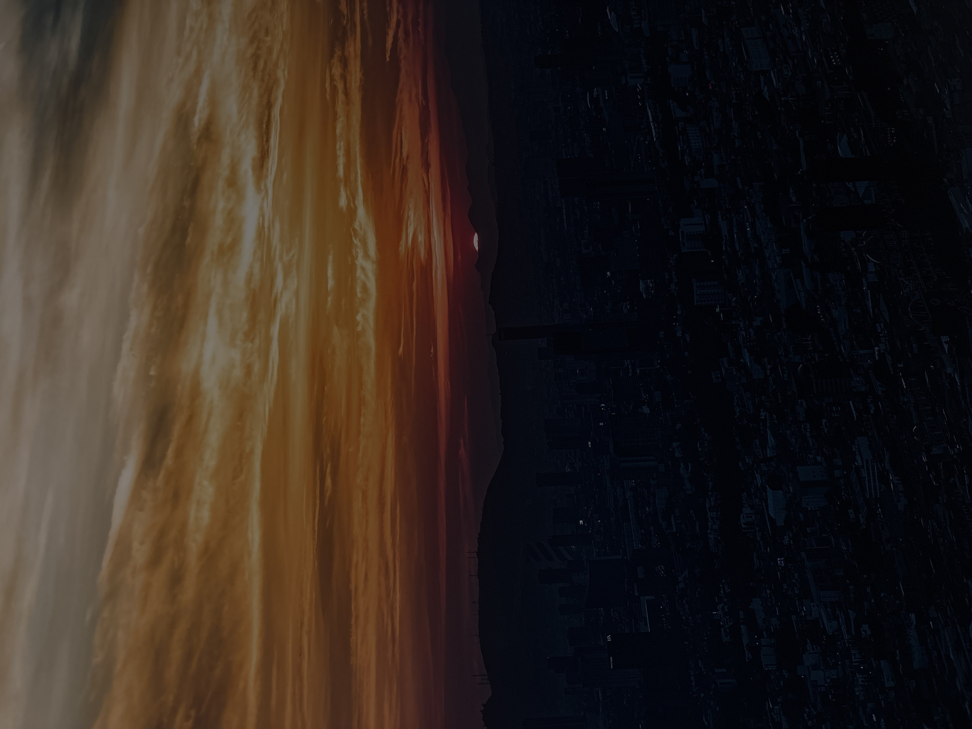 The image presents a dramatic view of a cityscape at either sunrise or sunset, set against a vibrant, streaked sky. The original image is oriented vertically, but it depicts a horizontal landscape.
The upper portion of the scene (left side in the original orientation) is dominated by a breathtaking sky, filled with long, wispy clouds illuminated by warm light. Fiery shades of orange, deep red, and yellow blend across the firmament, suggesting the intense colors of dawn or dusk. Some areas transition into softer, lighter hues, creating a sense of depth and atmospheric perspective.
Along the horizon line, which cuts across the middle of the image, a distinct silhouette of distant landforms and urban structures is visible. A small, reddish-orange sun disc peeks just above this horizon, indicating the precise moment of sunrise or sunset.
Below the horizon (right side in the original orientation), a sprawling cityscape is rendered in deep shadow. Buildings are dark, geometric silhouettes, creating a complex, almost abstract pattern of structures. Very few artificial lights are visible within the urban expanse, emphasizing the twilight conditions. The intricate details of the buildings are obscured by darkness, giving the city a monumental, yet mysterious, presence.
The overall impression is one of a vast, tranquil urban landscape captured during a transitional atmospheric event in Puebla, Mexico. Faint, almost indiscernible white alphanumeric characters, such as "VI" and "IA," can be observed on some of the dark building facades.