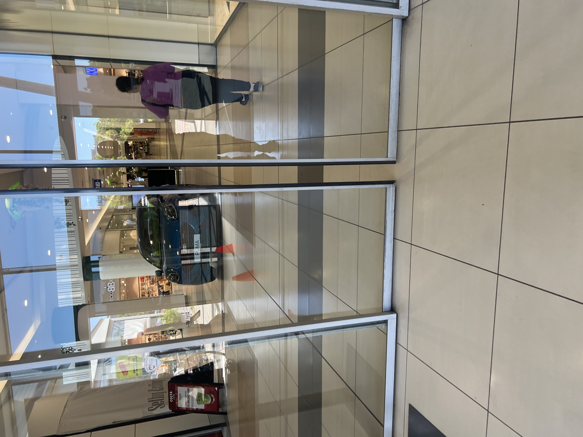 The image depicts a reflection in a glass storefront of a retail space, likely a shopping mall, located in Burgas, Bulgaria. Reflected within the glass are a dark-colored Ford Puma vehicle, promotional displays for Costa Coffee, and portions of other store interiors. Also visible is a person wearing a purple shirt walking through the mall. The flooring outside the storefront consists of light beige square tiles with dark grout lines.