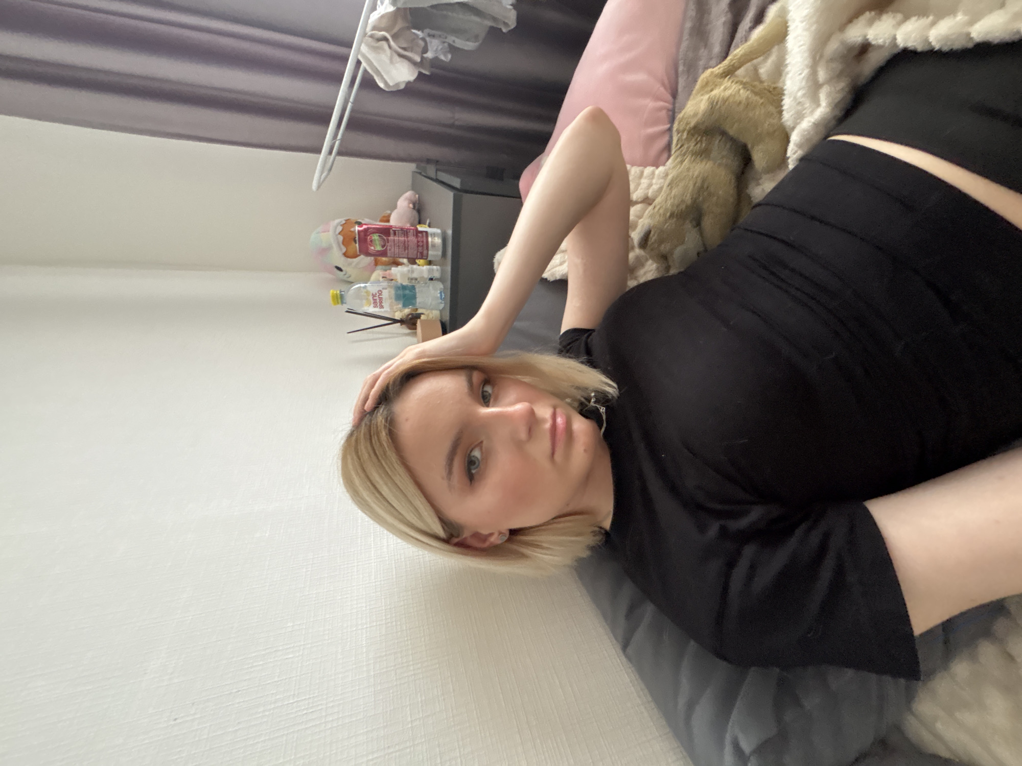 A blonde woman in a black shirt and shorts lies on a grey bed, her head near a white wall. A bedside table with water bottles, a toy, and other items is visible behind her head. A pink pillow, a tan stuffed animal, and a white blanket are on the bed near her. The scene is likely indoors, and a purple curtain is visible in the background. There is no verifiable information to confirm the location is Vsevolozhsk, Russia.