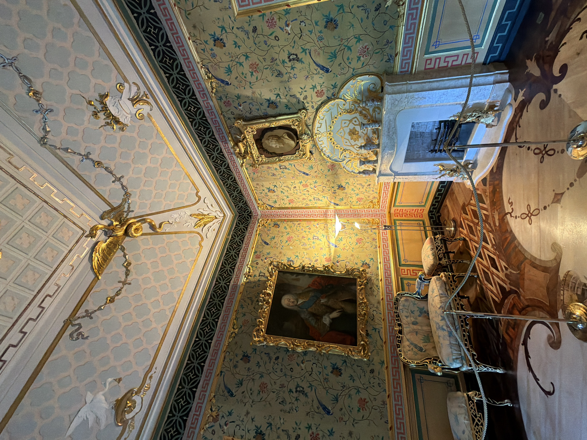 The image depicts an opulent room with ornately decorated walls, ceiling, and floor. The walls are covered in a floral wallpaper with birds and butterflies, framed by gold trim and decorative borders. A large framed portrait of a man in historical attire hangs on the left wall, while a smaller framed bust is mounted above a marble fireplace on the right wall. The fireplace has a decorative gilded mantelpiece.
The ceiling is intricately designed with a repeating geometric pattern in pale blue and white, accented with gold ornamentation, including a golden eagle-like figure and a decorative chain. The floor is made of polished wood with an inlaid geometric and swirling pattern.
In the foreground, there is a sofa with a matching stool, both upholstered in fabric with a similar floral design to the wallpaper, and featuring gilded wood frames. Two smaller stools or ottomans are positioned to the left of the sofa. A floor lamp stands behind the sofa. The room is cordoned off with gold rope and stanchions, indicating it is likely a museum or historical exhibit. The overall impression is one of extreme luxury and detailed craftsmanship. The location is identified as Unknown, Unknown.