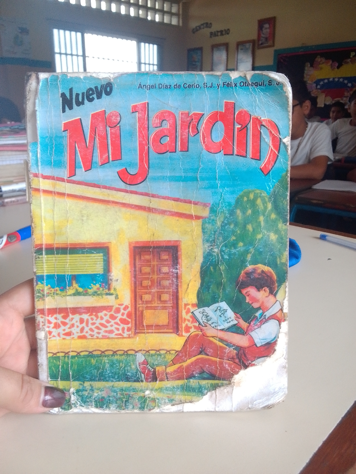 This is a photograph of a well-worn book cover featuring a vibrant illustration. The cover is mostly blue and yellow, with bold red lettering that reads "Nuevo Mi Jardin" at the top. Below the title, a yellow house with a red-patterned base and a blue window with yellow blinds is depicted. To the right of the house, a green tree stands behind a young boy sitting on a green lawn. The boy, wearing a blue shirt and a red vest, is engrossed in reading a book that shows pictures of a rabbit and a bird. The book cover shows signs of age and wear, with creases and tears visible throughout. In the background, glimpses of a classroom setting can be seen, including a window with horizontal blinds, framed pictures on the wall, and other students seated at desks. The overall impression is of an educational setting, possibly a school in Santa Ana, Venezuela, judging by the flag visible in the background. The lighting suggests it is daytime, with sunlight streaming through the windows.