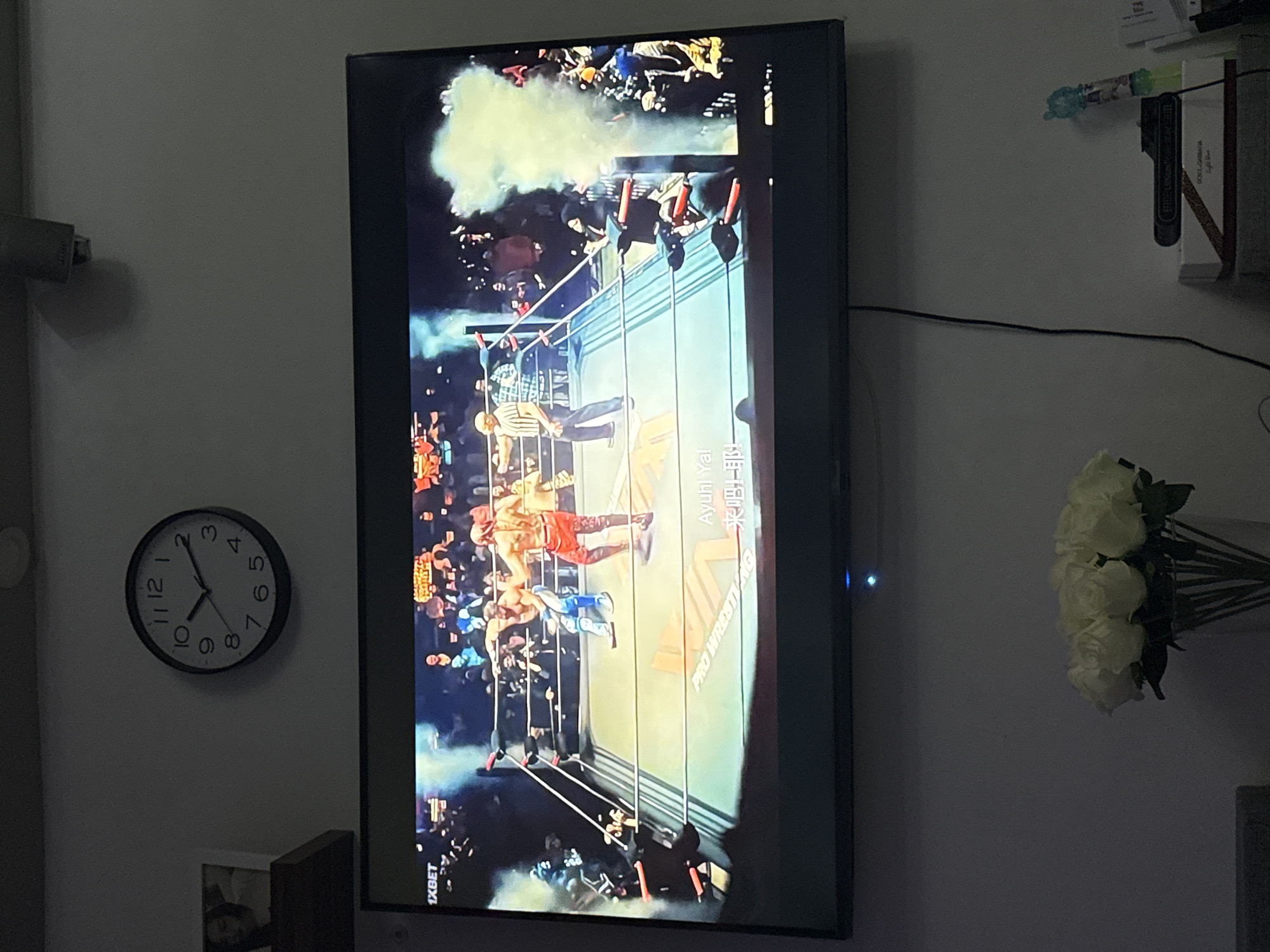 The image captures a television screen displaying a professional wrestling match. Two wrestlers are in the ring, with a referee standing nearby. Smoke billows into the arena from the side, adding to the dramatic atmosphere. The television is mounted on a wall above a vase of white roses. To the left of the television, a framed picture is visible on a shelf. A black wall clock with white numbers and hands is mounted above the television. The scene appears to be a living room or entertainment area in a home, potentially in Bat Yam, Israel, given the context provided.