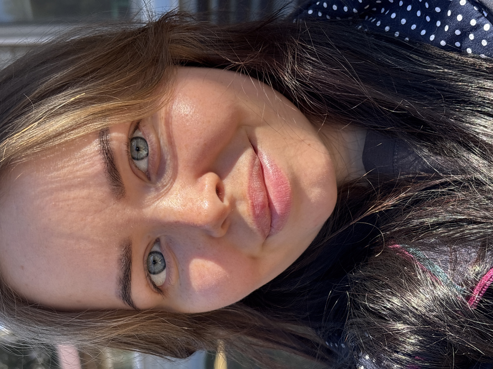 A close-up photograph shows a woman's face. She has fair skin, light green eyes, and brown hair with lighter brown highlights. She is wearing a dark shirt with white polka dots on the visible portion of the collar and sleeves. Part of a pink and teal braided strap can be seen in her hair. The environment or location is not visible. No further details can be deduced from the image.