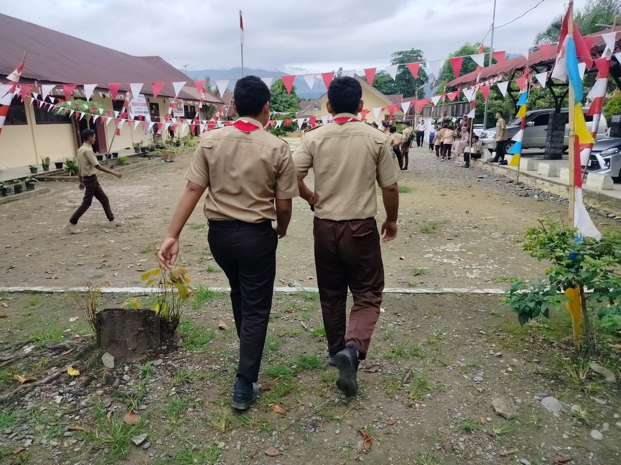 Two young males, dressed in scout uniforms consisting of tan shirts and brown trousers, walk away from the camera. They are in an outdoor setting, possibly a school or community event, with a building visible in the background adorned with red and white triangular flags. The ground is a mix of dirt and gravel, with some patches of grass. Several other people, also in similar uniforms or casual attire, are scattered throughout the scene. A flagpole with the Indonesian flag is visible in the distance. The overall atmosphere suggests a daytime event with a festive or ceremonial character. This scene is likely located in the city of Purwodadi, Indonesia, given the Indonesian flag and the commonality of scout activities in educational institutions there.