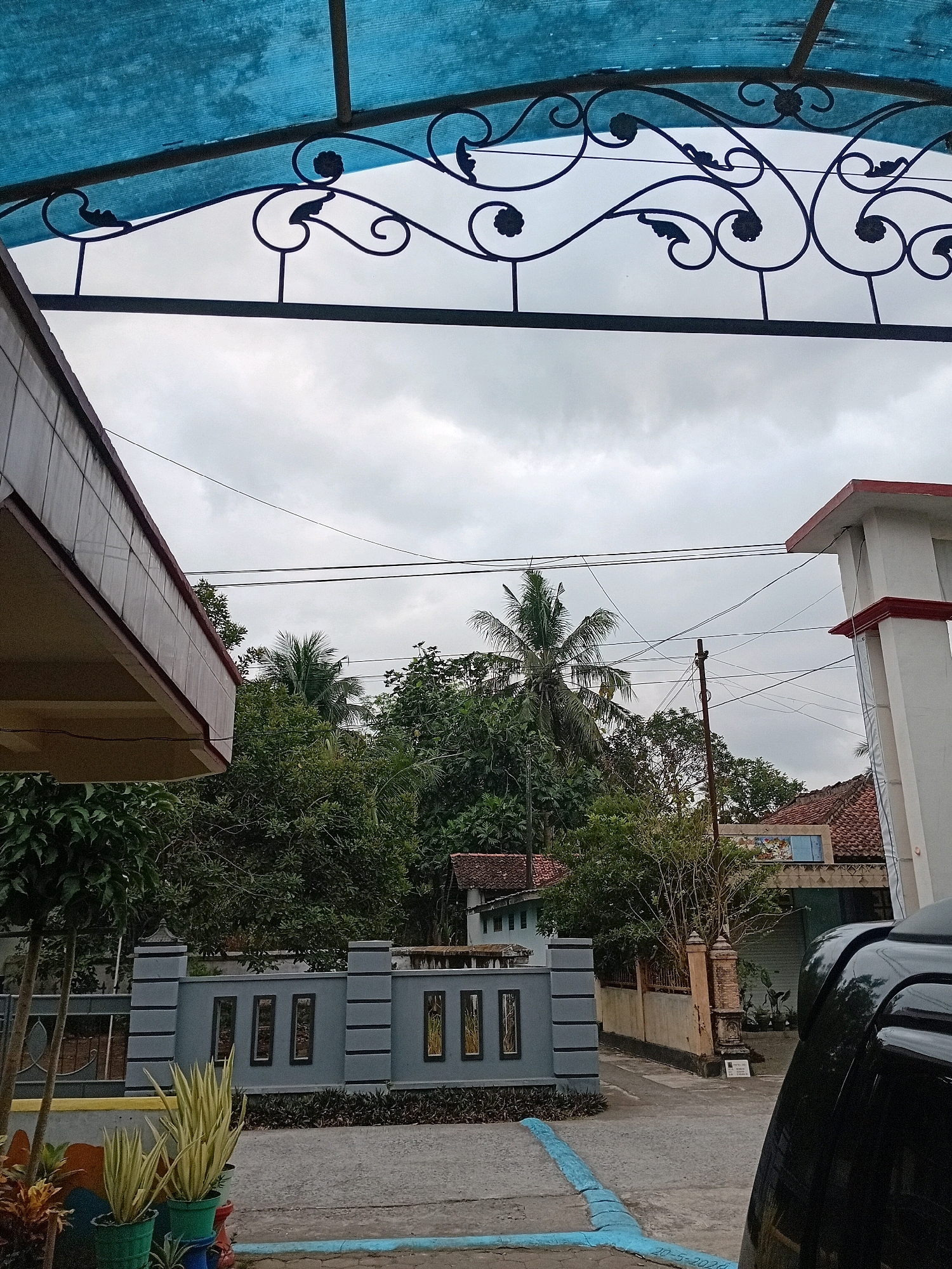 The image depicts an outdoor scene on an overcast day in Gebang, Indonesia. In the foreground, a black vehicle is partially visible on the right side, with its rear end pointing towards the right edge of the frame. A grey decorative fence with rectangular recessed panels runs horizontally across the middle ground. In front of the fence, two potted plants with long, slender, yellowish-green leaves are placed on the ground. To the left of the fence, a portion of a building with a tiled facade and a deep overhang is visible. Beyond the fence, dense green foliage from trees, including a prominent palm tree, fills the background. A white structure with red trim, resembling a gate or part of a building, is situated on the right side, with multiple power lines crisscrossing the sky above. The sky is covered in heavy, grey clouds.