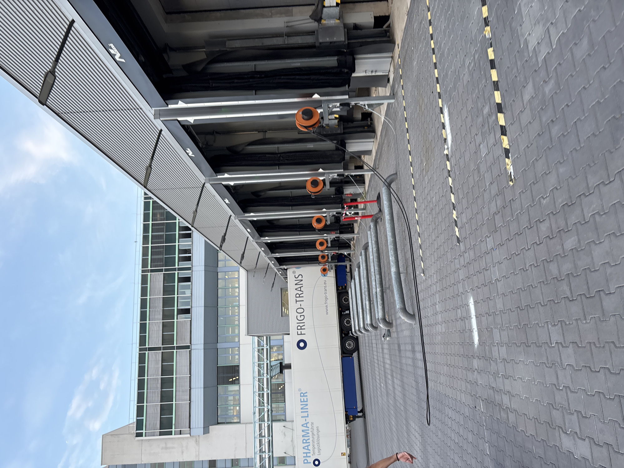 This is a photograph taken outdoors, possibly in Mannheim, Germany, during daylight hours under a partly cloudy sky. The scene depicts the loading docks of a commercial building. In the foreground, a grey paved area with interlocking bricks is visible, marked with yellow and black striped lines and some white paint. Several curved metal pipes run across the ground, connected to black cables.
To the left, a white semi-trailer truck with "PHARMA-LINER" and "FRIGO-TRANS" logos is parked. The truck is labeled with "Temperaturgeführte Logistiklösungen" and a website address.
The background features a modern building with large windows and a ribbed metal roof over the loading bays. Several orange cable reels are mounted on the walls near the loading docks. The overall environment suggests a logistics or distribution center. There is one person partially visible on the far left, wearing a black shirt and showing a hand.