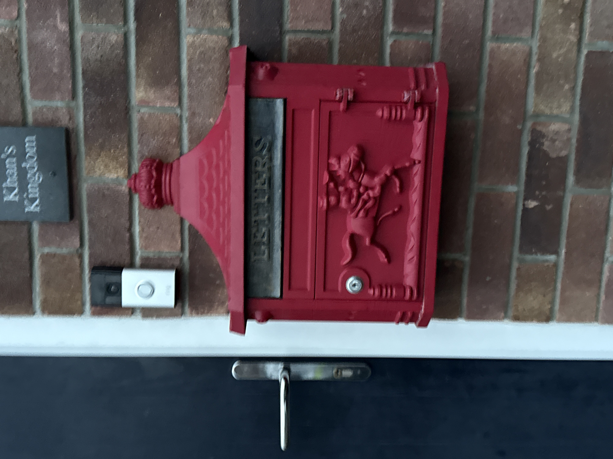 A close-up image shows the exterior of a house. A red mailbox with the word "LETTERS" displayed is affixed to a brick wall. To the left of the mailbox is a doorbell and a small plaque that reads "Khan's Kingdom". Below the mailbox and doorbell, a blue door with a silver handle is visible. The house is located in Great Yarmouth, United Kingdom.