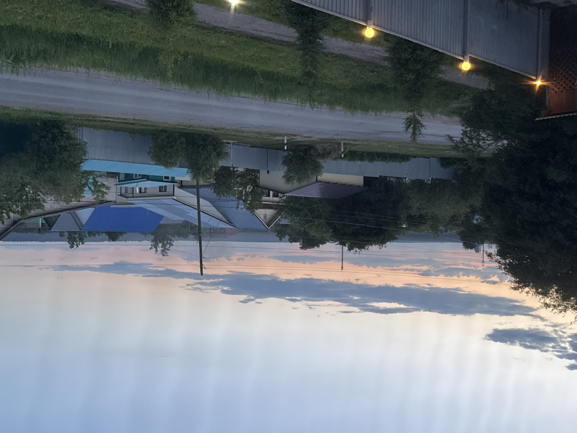 The image, correctly oriented in landscape, depicts a tranquil rural or semi-rural scene at twilight, likely either sunset or sunrise. The setting appears to be a village or small town, consistent with locations like Mozhga, Russia.
In the foreground, a gray road or dirt track runs horizontally across the frame, bordered by green grass and scattered trees. Along the right side of the image, a corrugated metal fence features several spherical, glowing yellow lights, and another brighter light source is visible further right, indicating artificial illumination during low light conditions.
Beyond the road and trees, a collection of houses with diverse roof colors, including blue, light blue, and grey, are visible. Utility poles and overhead wires crisscross the mid-ground, suggesting a modest infrastructure. Further in the background, a wide body of water, possibly a lake or a broad river, stretches across the landscape, reflecting the soft hues of the sky. The distant shore across the water is lined with trees and faint landmasses.
The sky dominates the upper half of the image, displaying a gradient of colors from soft blue at the top, transitioning through pale pinks and oranges towards the horizon. Elongated clouds, tinged with these warm and cool colors, add to the atmospheric quality of the scene.
There are no people or specific activities taking place, contributing to a sense of calm and quiet. The notable details include the glowing lights, the blend of natural and artificial light, and the serene color palette of the sky, all indicative of a peaceful twilight moment. No visible text is present in the image.