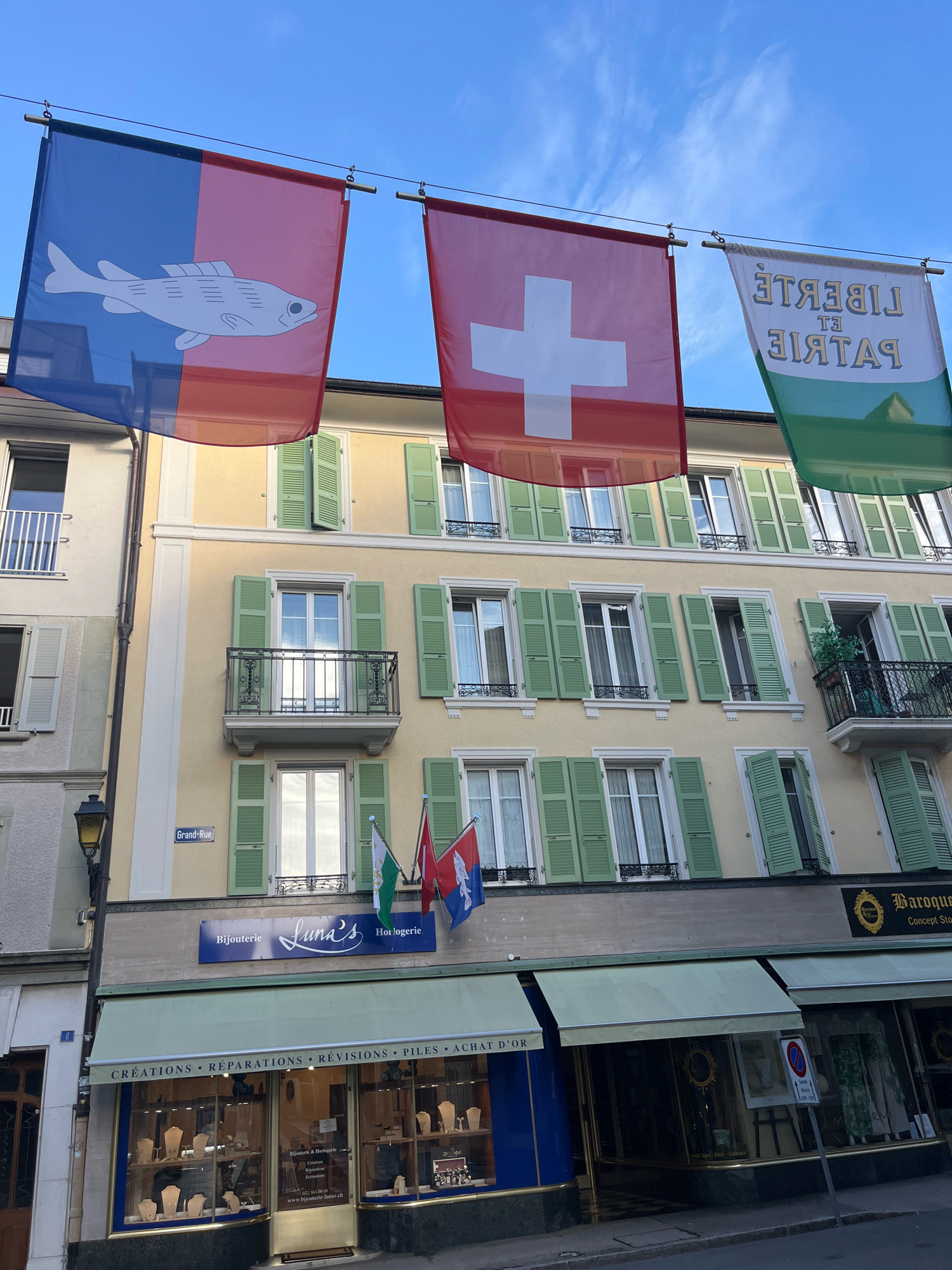 The image shows a street scene in Nyon, Switzerland, with flags hanging across the street in front of a building. The building is a light yellow, with green shutters on the windows. The flags are of Switzerland, and two other flags, one with a fish and one with the text "LIBERTE ET PATRIE". Below the flags, there is a storefront with the name "Bijouterie Luna's Horlogerie" and the words "CREATIONS REPARATIONS REVISIONS PILES ACHAT D'OR". There is a street sign that says "Grand-Rue". The sky is blue with some clouds. The image appears to have been taken during the daytime.