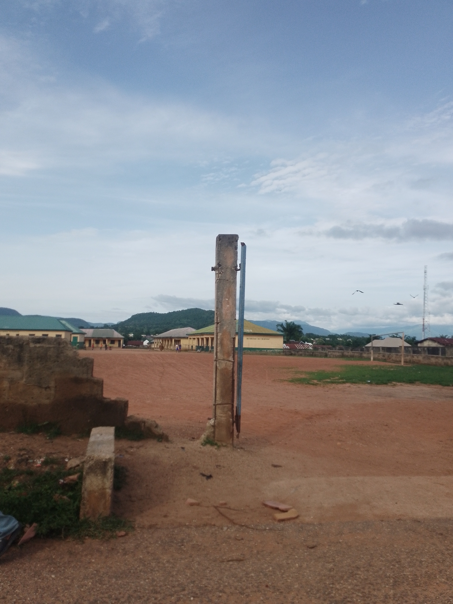 The image depicts an outdoor scene, likely a schoolyard or playing field, with a prominent concrete pillar in the foreground. The ground is covered in reddish-brown dirt, with some patchy grass and small stones. To the left, a rough-hewn concrete structure with visible steps is present. In the background, several buildings with green and yellow roofs are visible, interspersed with trees and hills. The sky is overcast with some clouds. Three birds are flying in the distance. A tall communication tower can be seen on the right side of the frame. There are no people clearly visible in the image.
