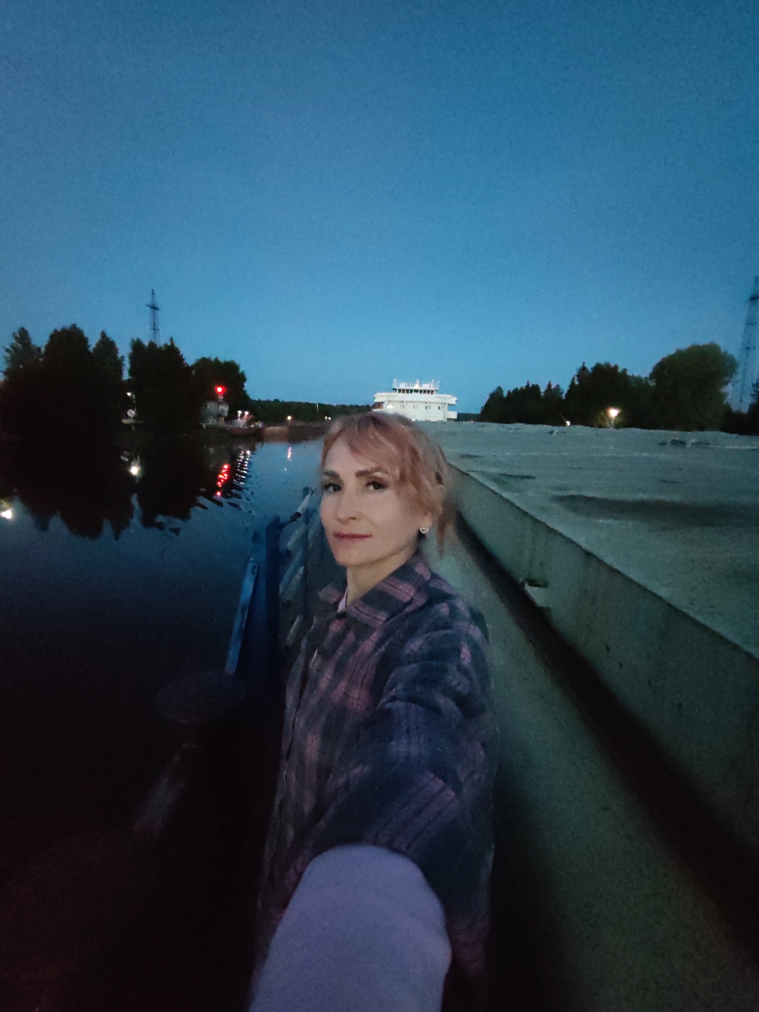 A woman with short, light brown hair stands on a paved surface next to a body of water at dusk. She is wearing a pink and blue plaid shirt and a light purple sleeve is visible in the lower right corner. She is looking towards the camera with a slight smile. In the background, across the water, are trees and a building resembling a ship. There are lights reflecting on the water. The sky is a deep blue. The scene appears to be located near Annensky Most, Russia, as indicated by the presence of a waterway and elements common to that region.