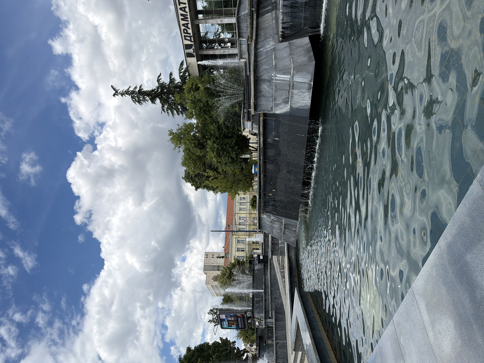 The image depicts the central fountain area in the city of Pazardzhik, Bulgaria. The fountain is a multi-tiered structure made of dark stone, with water cascading over its edges and spraying from several jets. In the foreground, the water is calm with gentle ripples reflecting the sky. Behind the fountain, there are trees and several buildings. To the right, a modern building with large glass windows bears Cyrillic text reading "ДРАМАТИЧЕН" (Dramatic), suggesting it is a drama theater. To the left, a billboard displays advertisements, and further back, there are more traditional buildings with red tiled roofs. The sky is blue with scattered white clouds, indicating a pleasant day. No people are clearly visible in the scene.