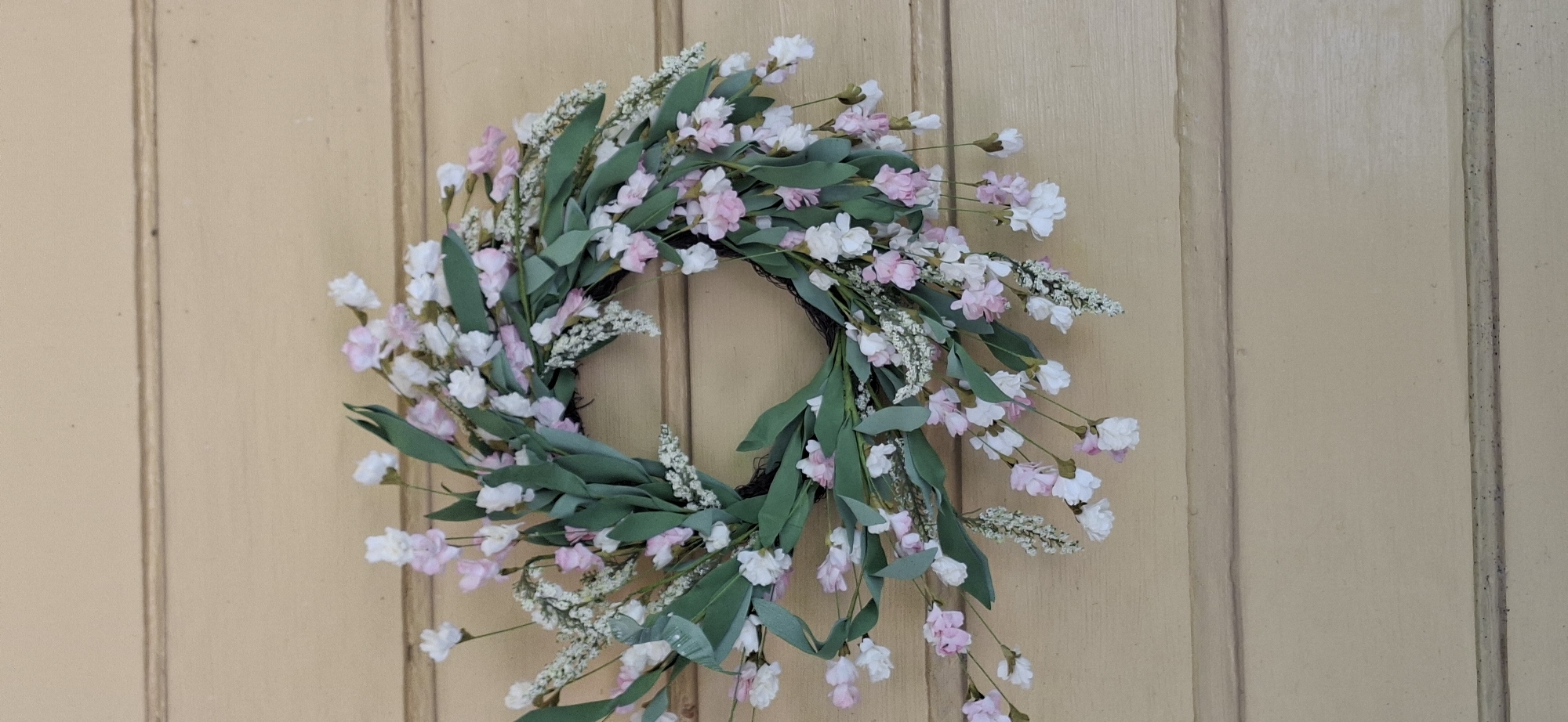 A wreath with pink and white flowers and green leaves is displayed on a light brown wooden surface. The wreath is circular and has a natural, organic appearance. The flowers are small and delicate, and the leaves are a deep green. The wooden surface appears to be made of horizontal planks, possibly a door or a wall. The lighting is even, and there are no shadows, suggesting an indoor setting or an overcast day. The image is a close-up of the wreath, with the background out of focus. The overall impression is one of rustic charm and natural beauty.