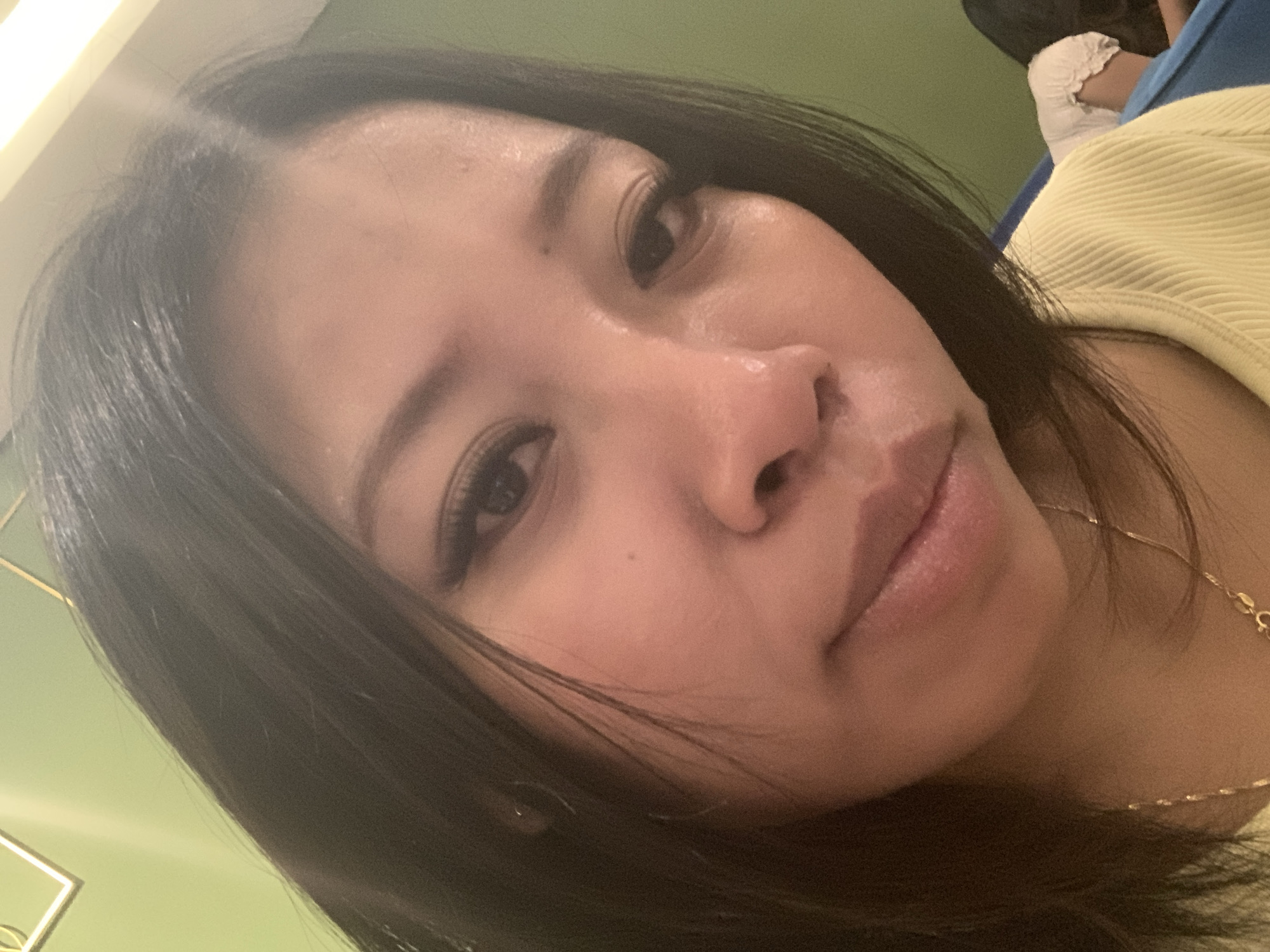 A close-up, indoor selfie features a young woman with dark hair and dark eyes, her face illuminated by soft lighting. She is wearing a light yellow ribbed top and a delicate gold necklace. The background consists of a light green wall with a gold geometric accent, suggesting an indoor setting possibly in Denpasar, Indonesia. The overall impression is informal and personal.