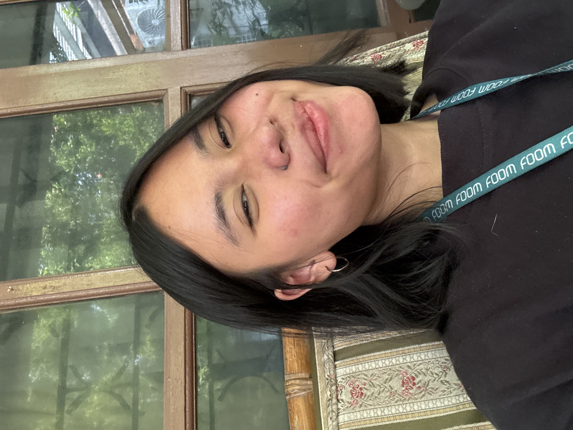 A young woman with dark hair and a light complexion is sitting on a patterned chair in front of a window. She is wearing a black crewneck sweatshirt. A teal lanyard with white text is visible around her neck. The window behind her has a wooden frame and shows greenery and an air conditioning unit. The scene appears to be an outdoor or semi-outdoor setting, possibly a cafe or residential area in South Jakarta, Indonesia, given the architectural elements and vegetation.
