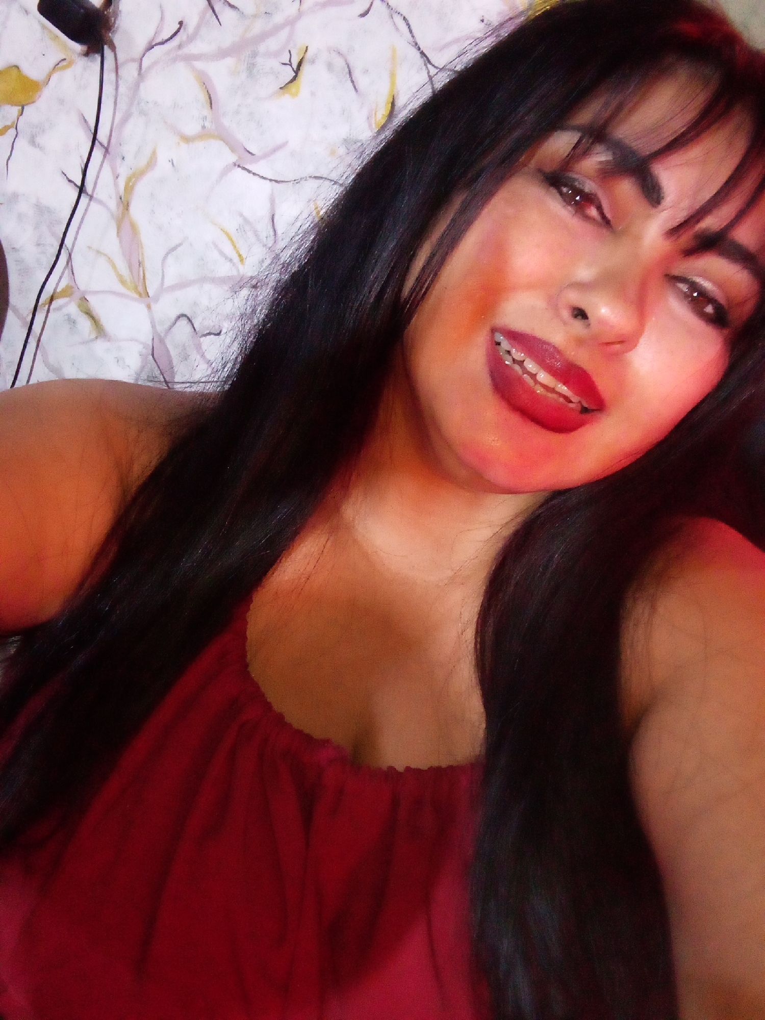 Here is a detailed description of the image:
A woman with long black hair and bangs is facing the camera with a slight smile. She is wearing a red top. Her lips are painted red, and she appears to have braces on her teeth. The background consists of a white surface with a branch-like pattern in light colors. Part of a black cord can be seen in the upper-left corner of the image. The woman appears to be the primary subject, and the focus is on her face and upper body. The location is not specified in the prompt.