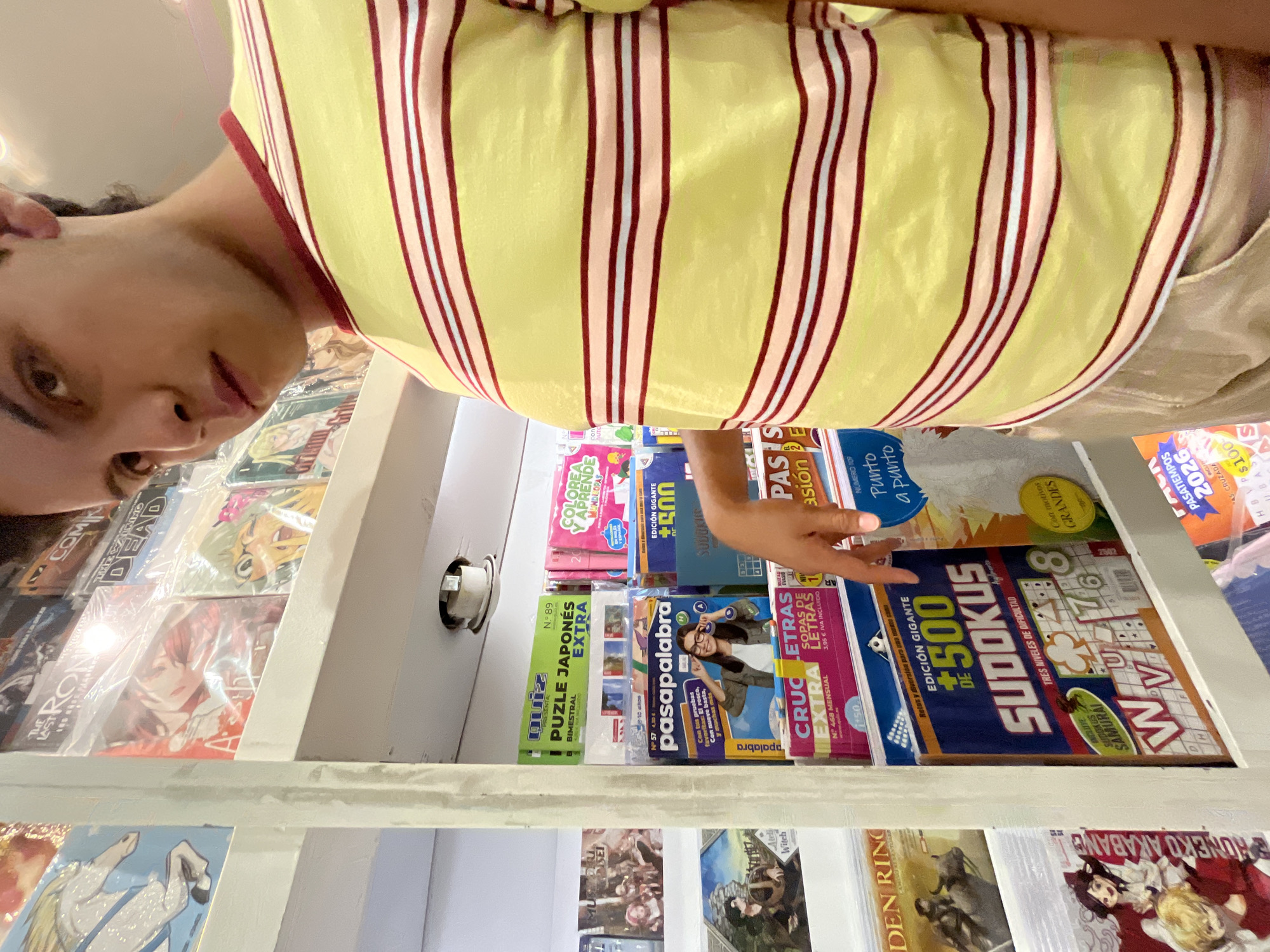 A person is looking at a display of puzzle and activity books in a store. The books include titles like "Sudokus", "Pasapalabra", and "Puzle Japones Extra". There are also manga volumes in the shelves above and below. The location could be in Puebla, Mexico based on the Spanish language books. The person is wearing a yellow striped shirt.