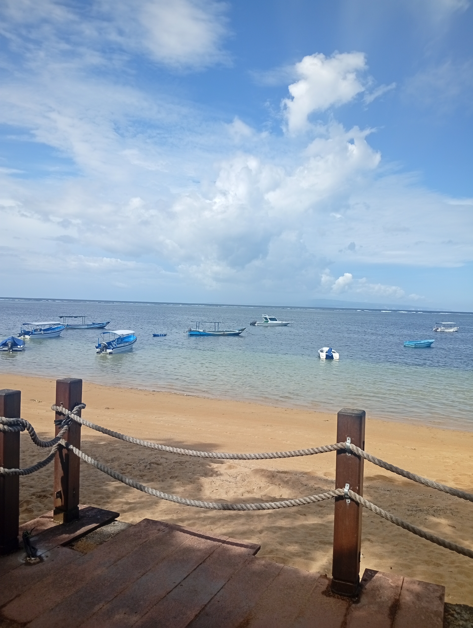 The image captures a coastal scene in Denpasar, Indonesia. The foreground features a weathered wooden dock with rope railings, transitioning to a sandy beach. Calm, shallow water extends to the horizon, dotted with various small boats, some with canopies. The sky is partly cloudy, with a gradient of blue and white hues. The overall lighting suggests a bright, sunny day. No people are visible in the frame.