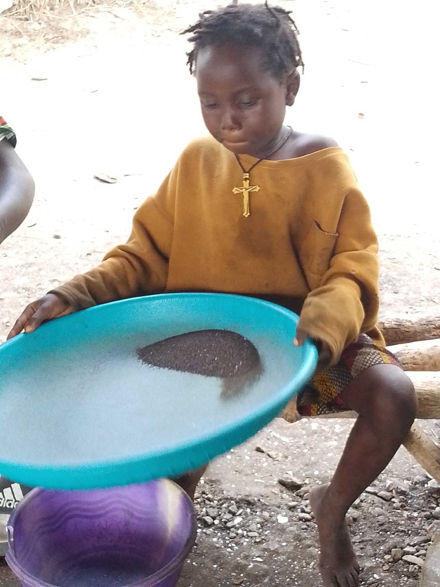 A young child, likely of African descent, is the sole focus of this media file. The child is seated, holding a bright blue plastic basin filled with what appears to be water and dirt. Their expression is one of concentration, with eyes cast downwards towards the basin. The child is wearing an oversized, mustard-yellow sweater that has been pulled off one shoulder, revealing their collarbone. Around their neck hangs a prominent gold cross pendant on a black cord. Below the sweater, a patterned skirt or shorts with a colorful, geometric design is visible. The child's bare feet are visible, and one leg is bent, while the other is extended.
The setting appears to be outdoors, with a sandy or dusty ground scattered with small stones and twigs. A rough wooden beam is visible behind the child, suggesting a rustic or natural environment. To the left, another person's arm, wearing a patterned garment, is partially visible, indicating the presence of at least one other individual. Beneath the blue basin, a purple plastic bucket is positioned, possibly to catch runoff or hold materials. The lighting suggests it could be daytime, but the specific time or weather conditions are not discernible. The overall impression is one of simple activity, possibly related to washing or sifting materials, set in a humble outdoor location. There is no visible text in the image.